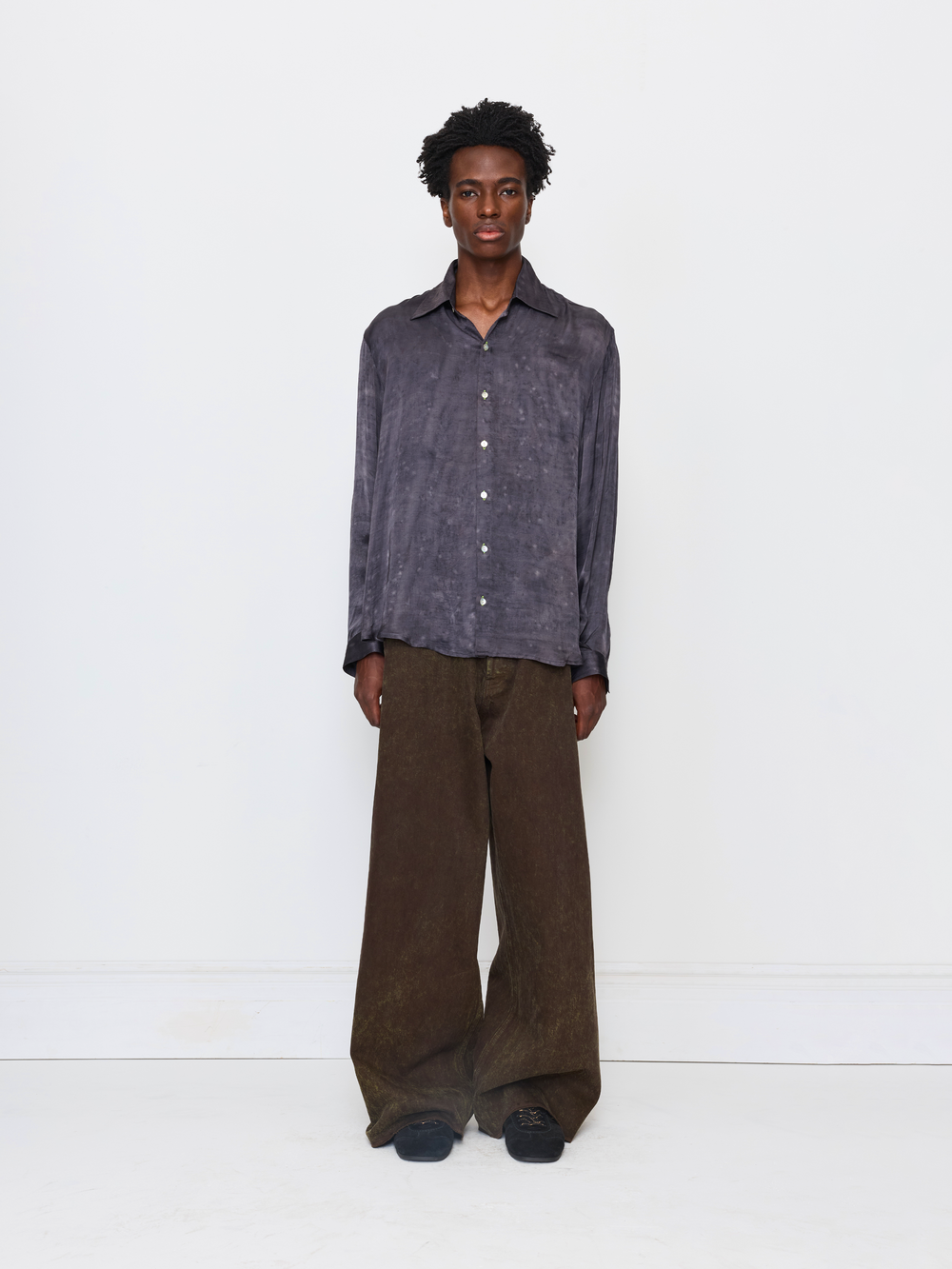 A model stands against a plain white studio background wearing a dark gray, textured long-sleeve button-up shirt and wide-leg brown trousers.