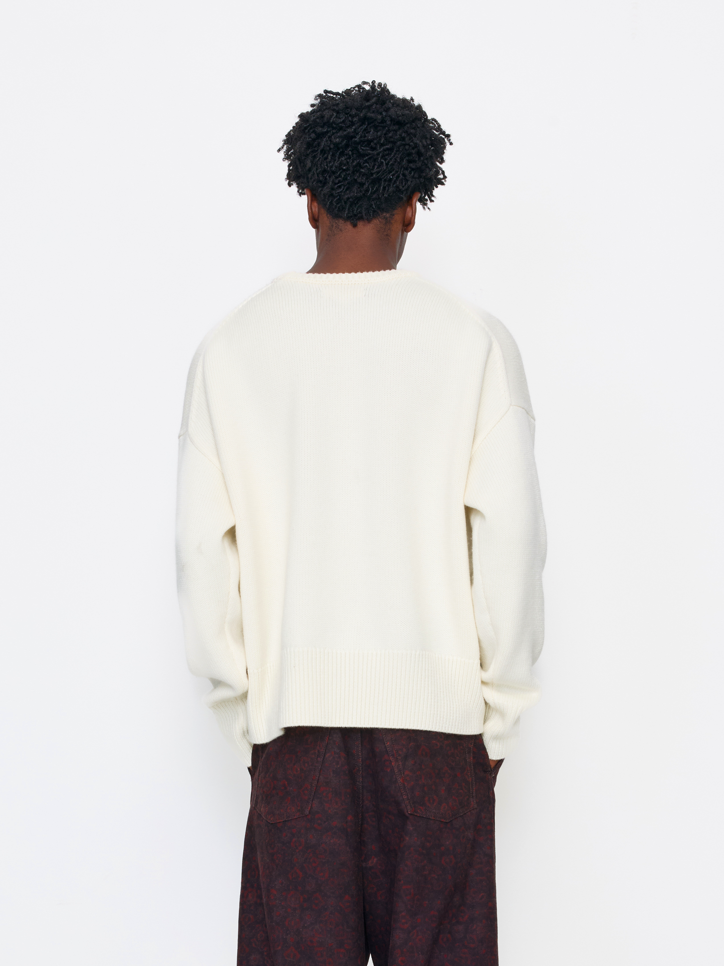 A person shown from the back wearing an oversized cream knit sweater and dark patterned trousers against a plain white studio background.
