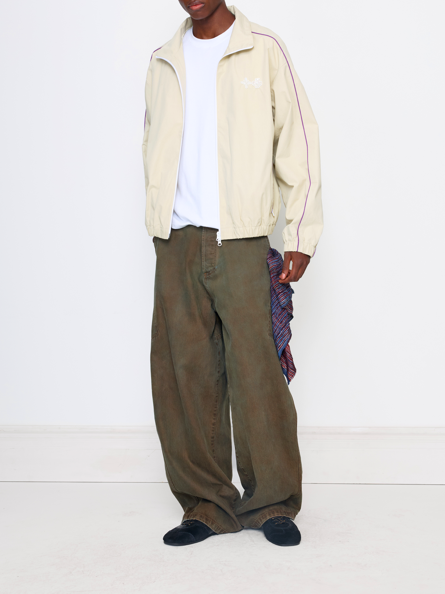 A model is shown from the neck down wearing a cream-colored track jacket with pink stripes, olive green wide-leg pants, a white t-shirt, and black shoes, holding a patterned scarf.