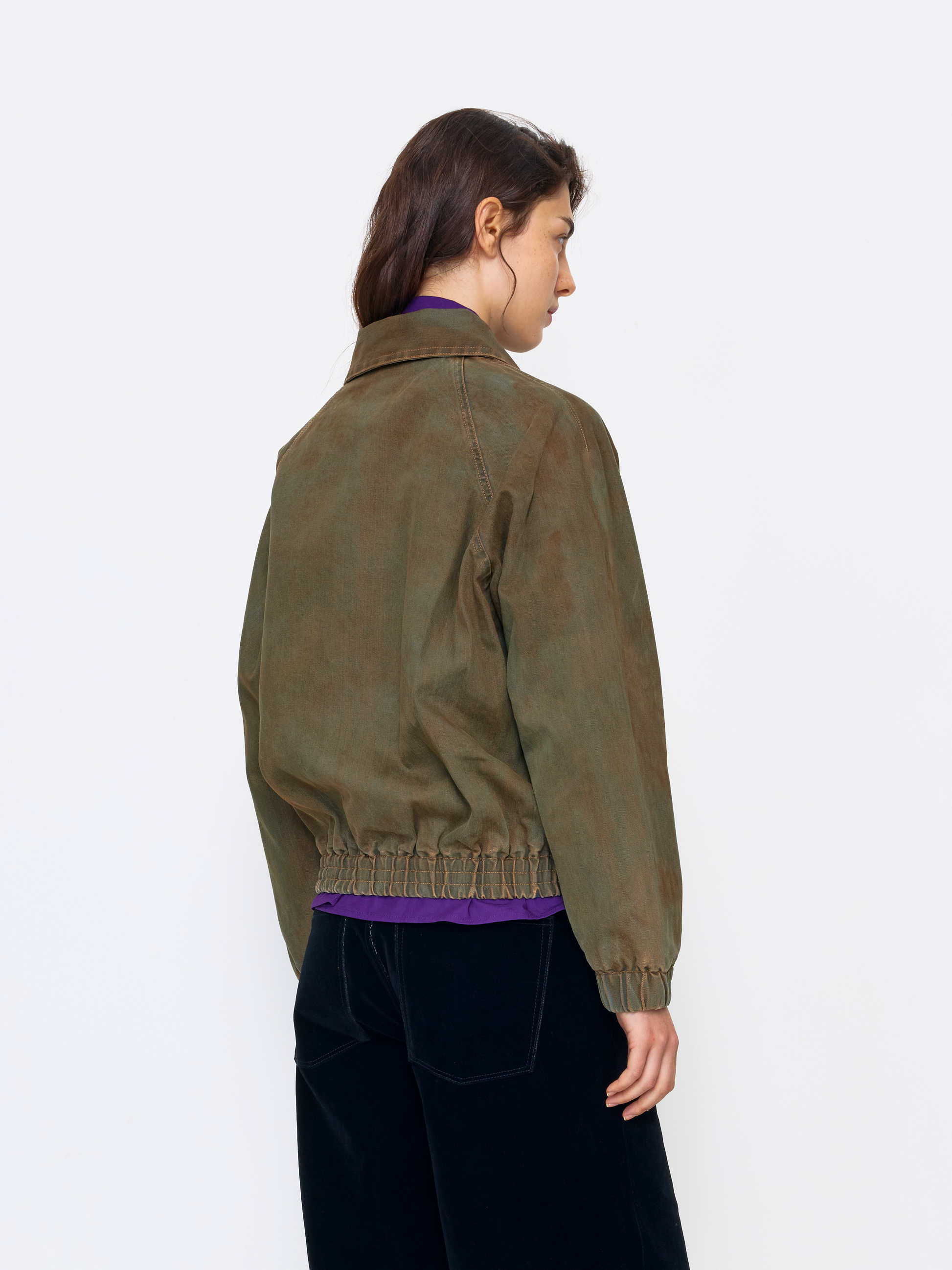 A model shown from the back wearing an olive green jacket with a gathered elastic hem, paired with black pants and a purple shirt underneath, against a plain white studio background.