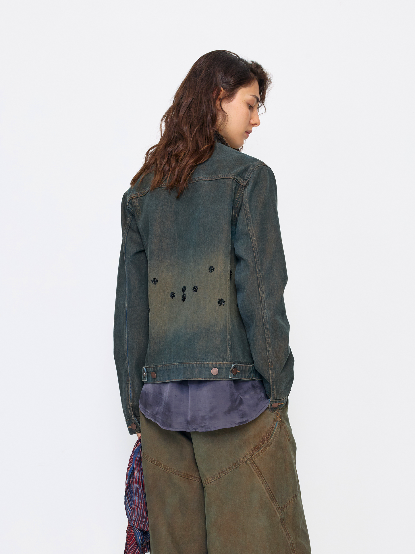 A model shown from the back wearing a dark washed denim jacket with small black embroidered details on the back, layered over a purple shirt and olive green pants, against a plain white background.