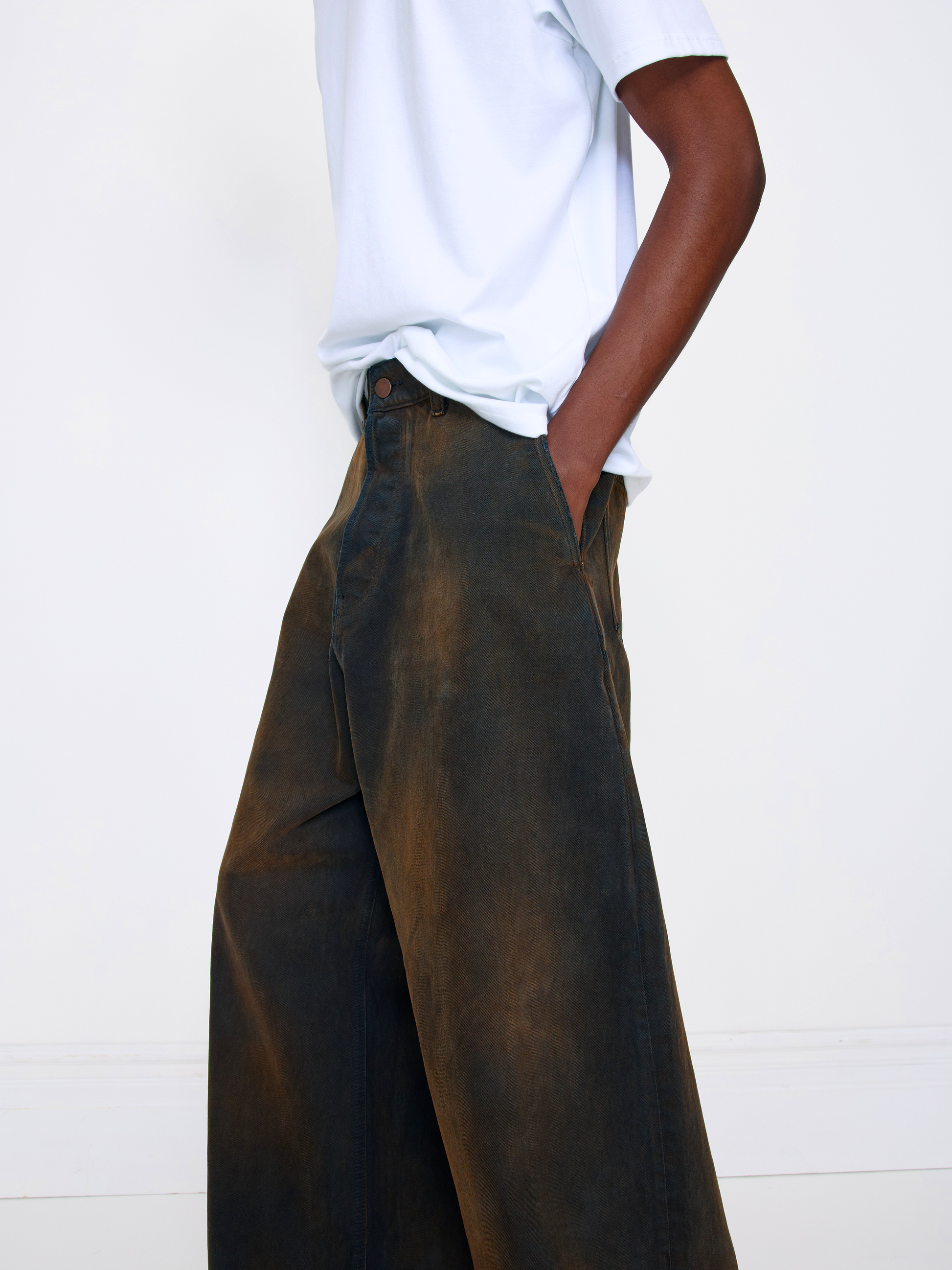 A person wearing wide-leg dark brown washed trousers and a white t-shirt, standing against a clean white wall.