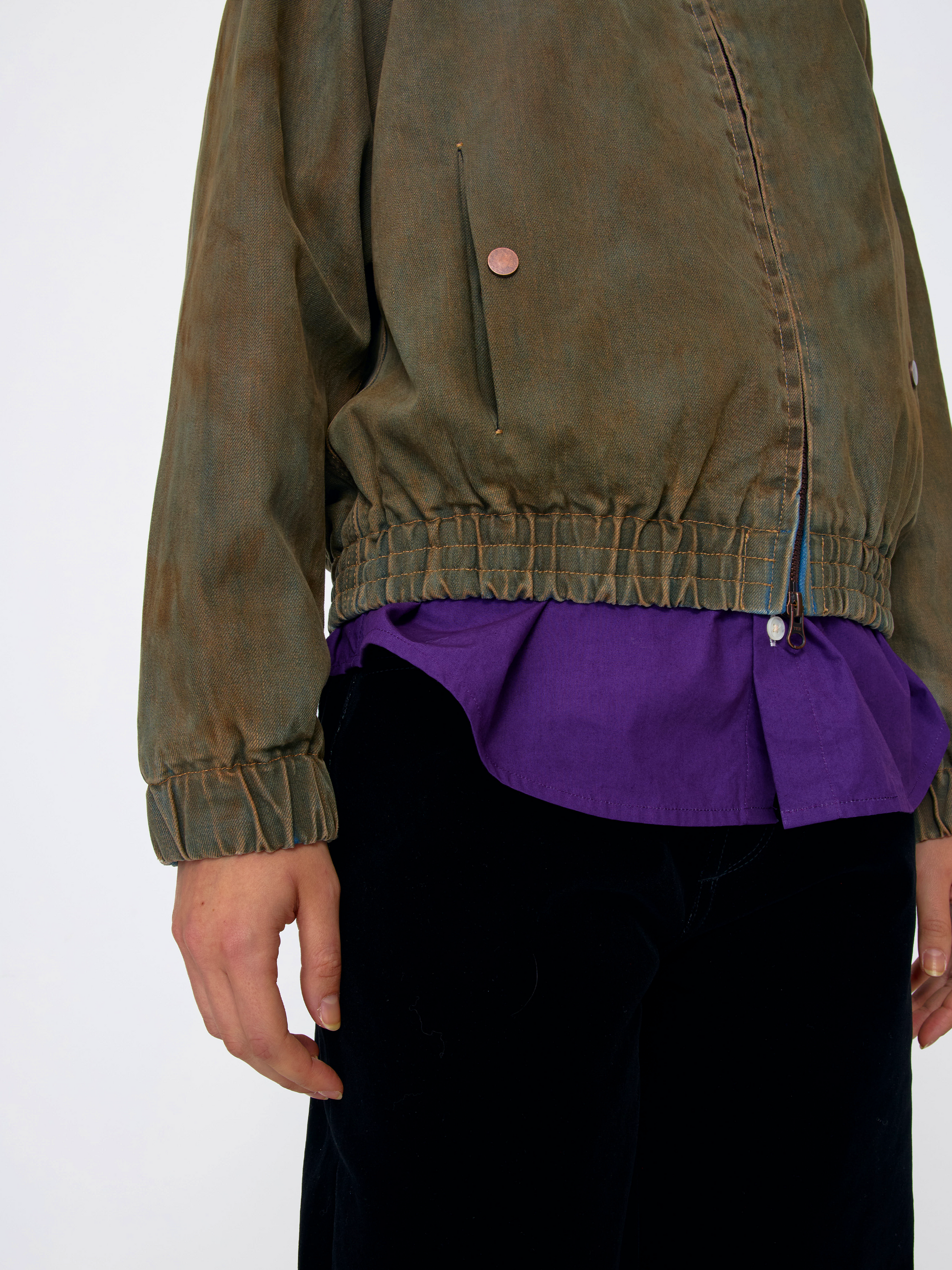 A cropped view of a person wearing an olive-brown jacket over a purple shirt and black pants, shown against a plain light background.