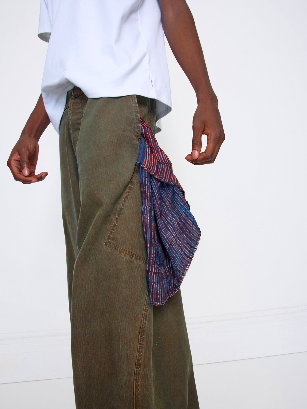 A person wearing wide-leg olive green pants with a striped red, blue, and purple fabric accessory draped over the side pocket, paired with a white t-shirt against a plain white wall.