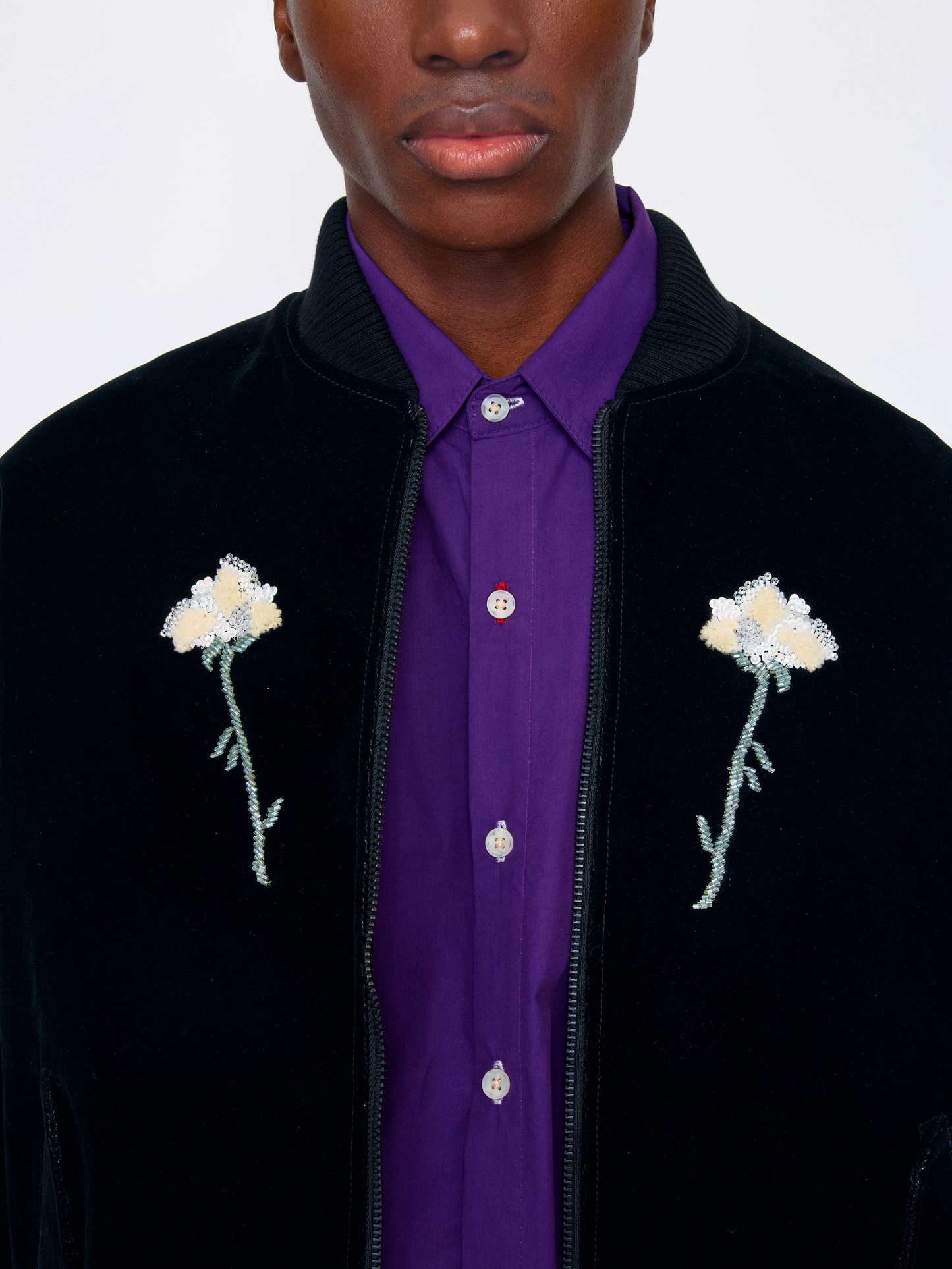 A close-up of a model wearing a black velvet bomber jacket with embroidered flowers on the chest, layered over a purple button-down shirt.