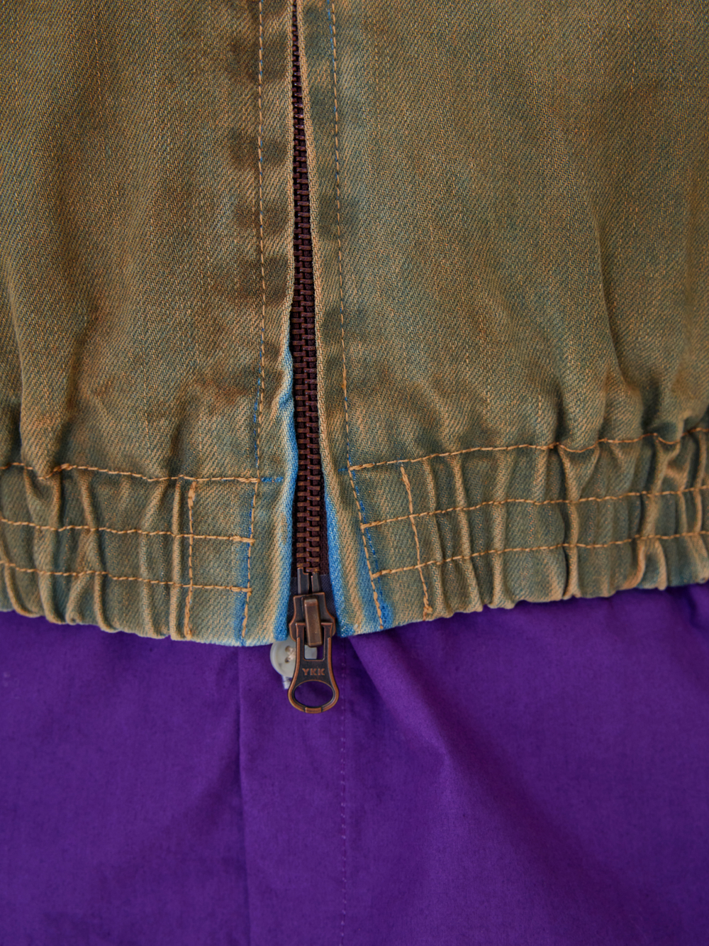 Close-up of a garment with an olive green fabric top section, elasticized hem, tan zipper, and a purple lower section.