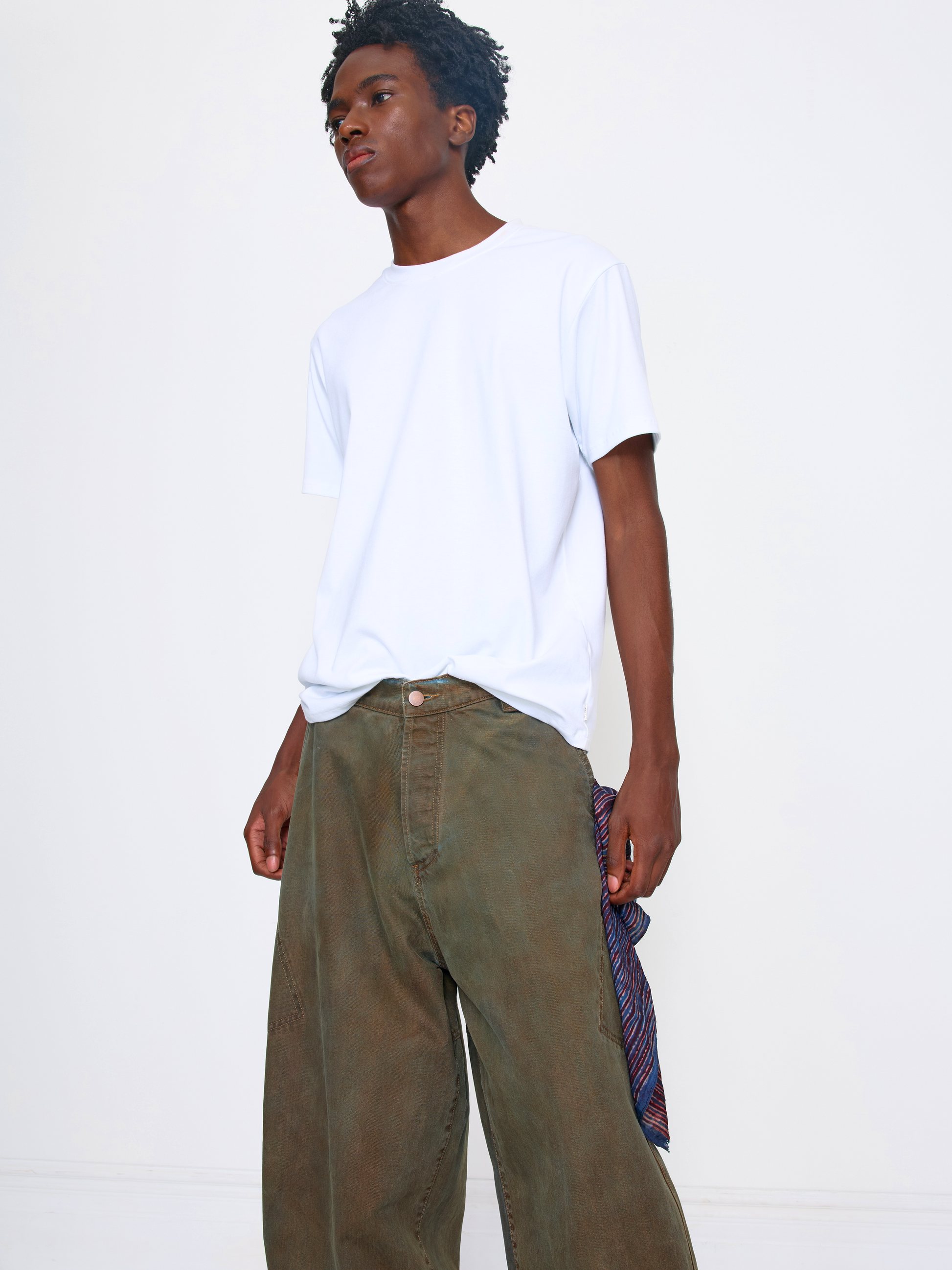 A model wearing a plain white short-sleeve crewneck t-shirt tucked into wide-leg olive green pants, holding a patterned scarf against a clean white studio background.