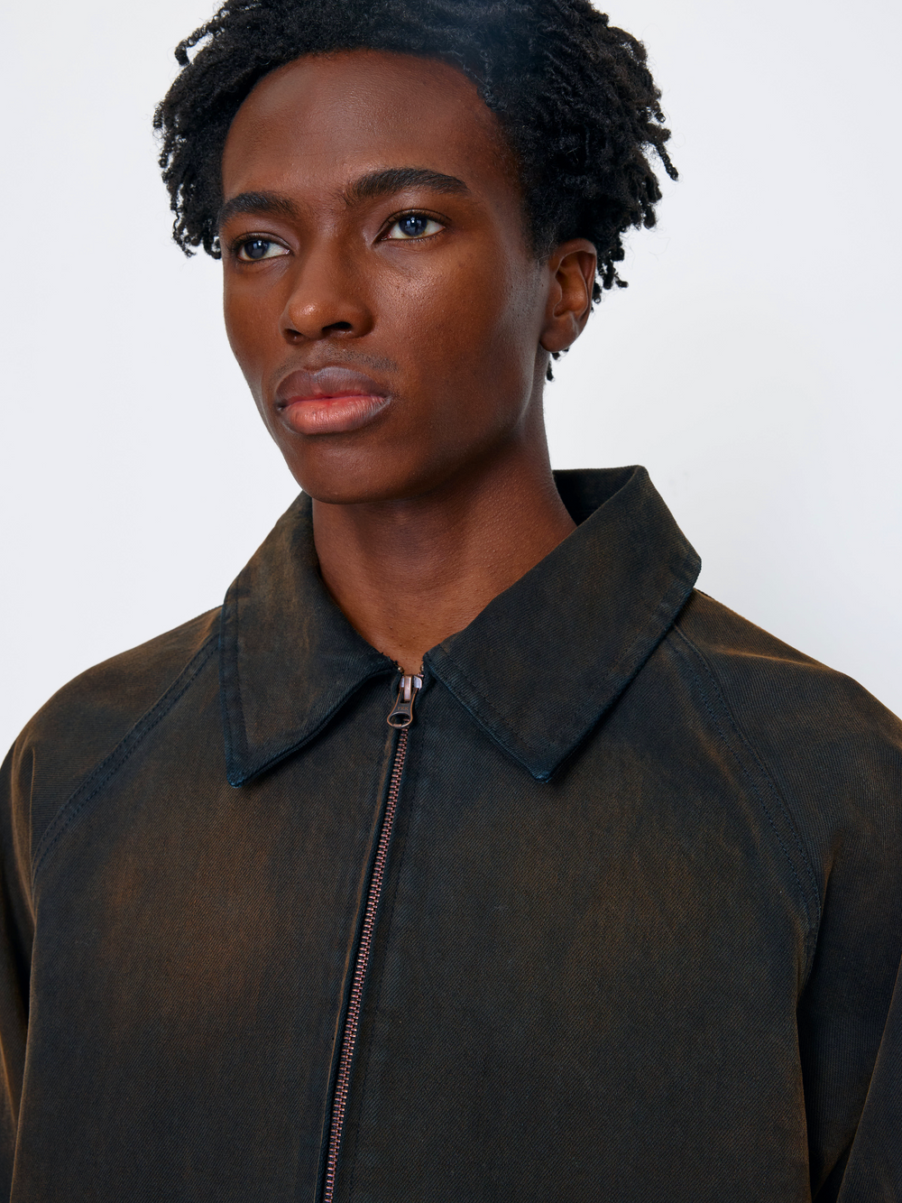 A close-up portrait of a model wearing a dark brown textured zip-up jacket with a wide collar, set against a plain light gray background.