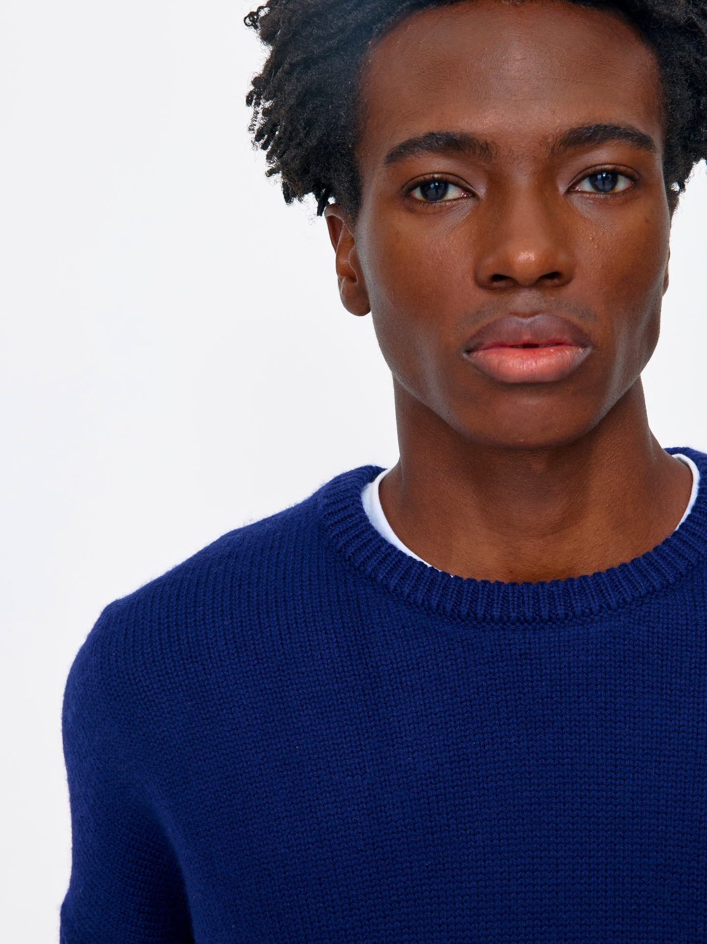 A close-up portrait of a man wearing a navy blue knit crewneck sweater over a white undershirt, set against a plain light gray background.