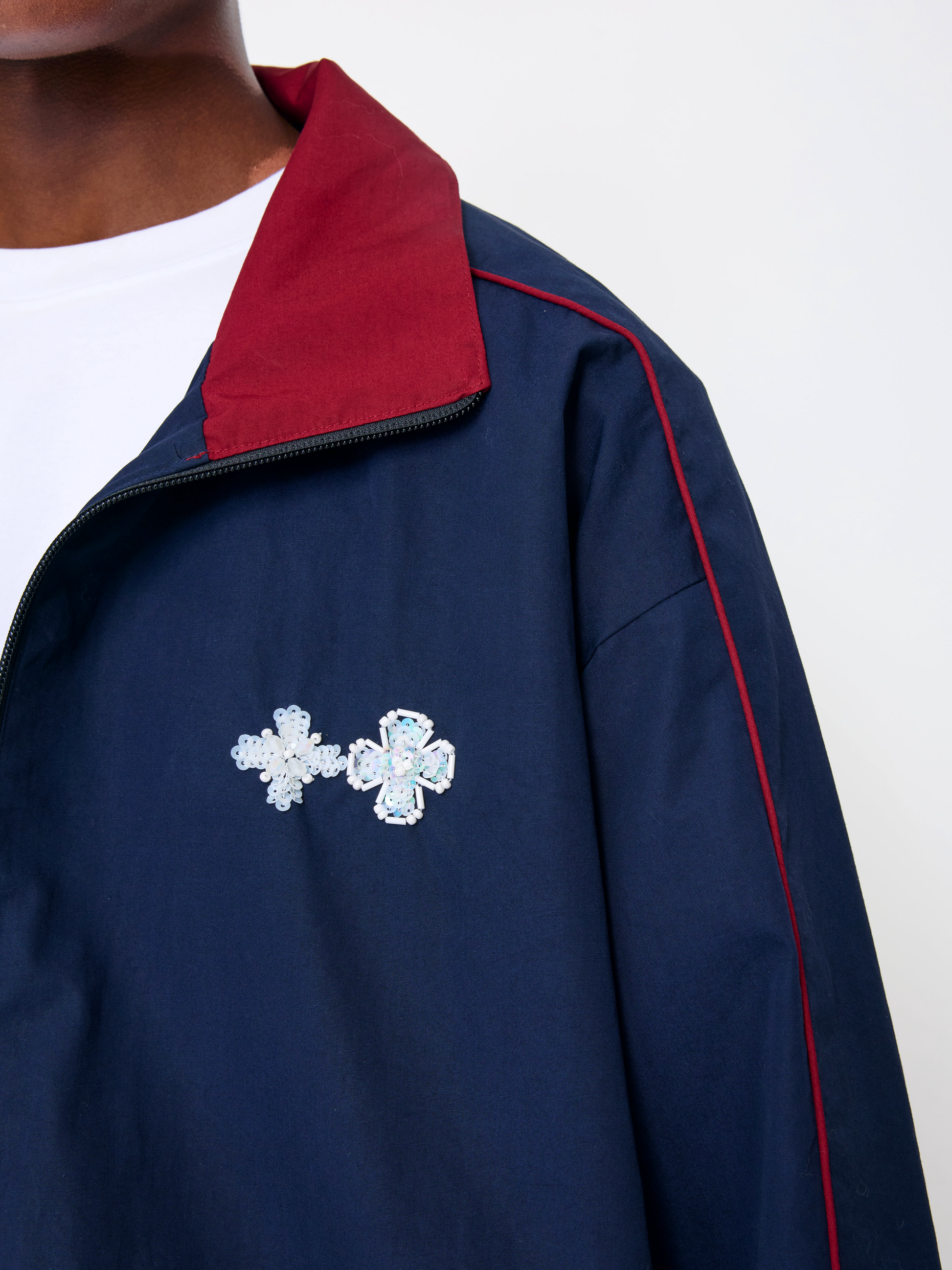 Close-up of a navy blue track jacket with a red collar, red piping, and two silver rhinestone cross brooch pins on the chest, worn over a white t-shirt.