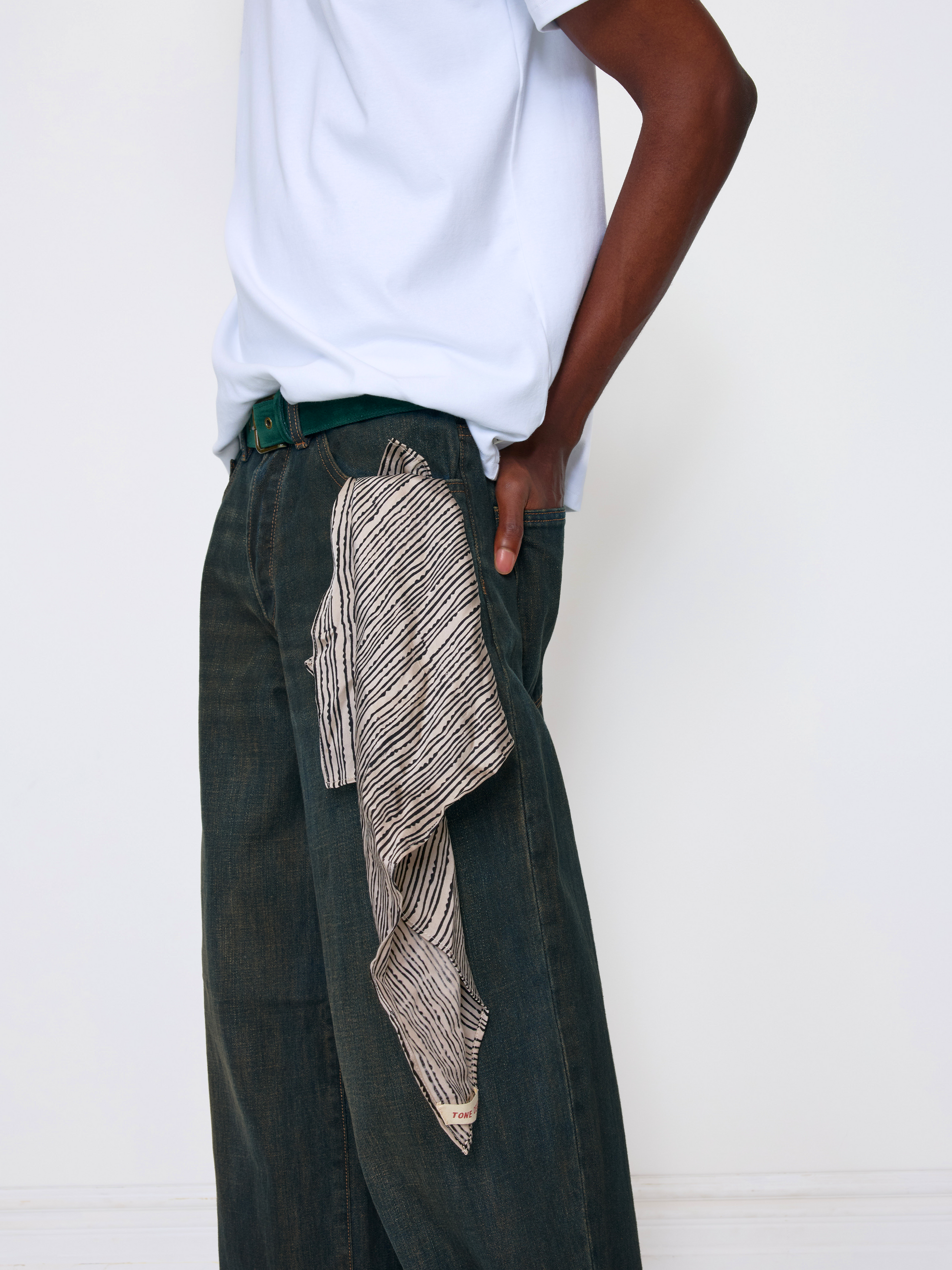A person wearing dark wide-leg jeans with a beige and black striped scarf draped from the side pocket, paired with a white t-shirt and a green belt, against a plain white wall.