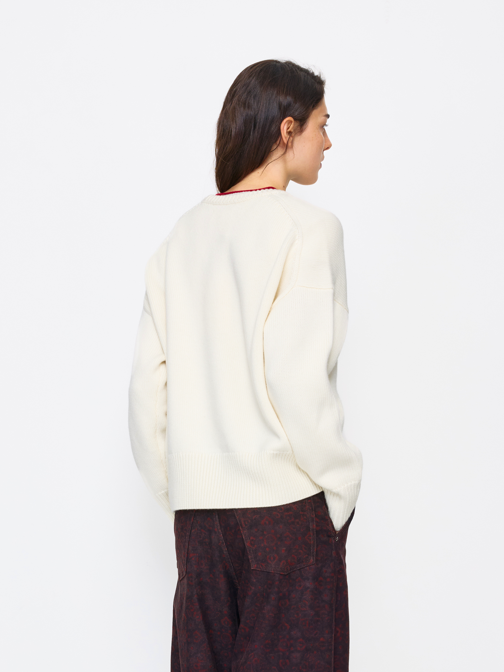 A woman shown from the back wearing a cream-colored knit sweater and dark patterned trousers, photographed against a clean white studio background.