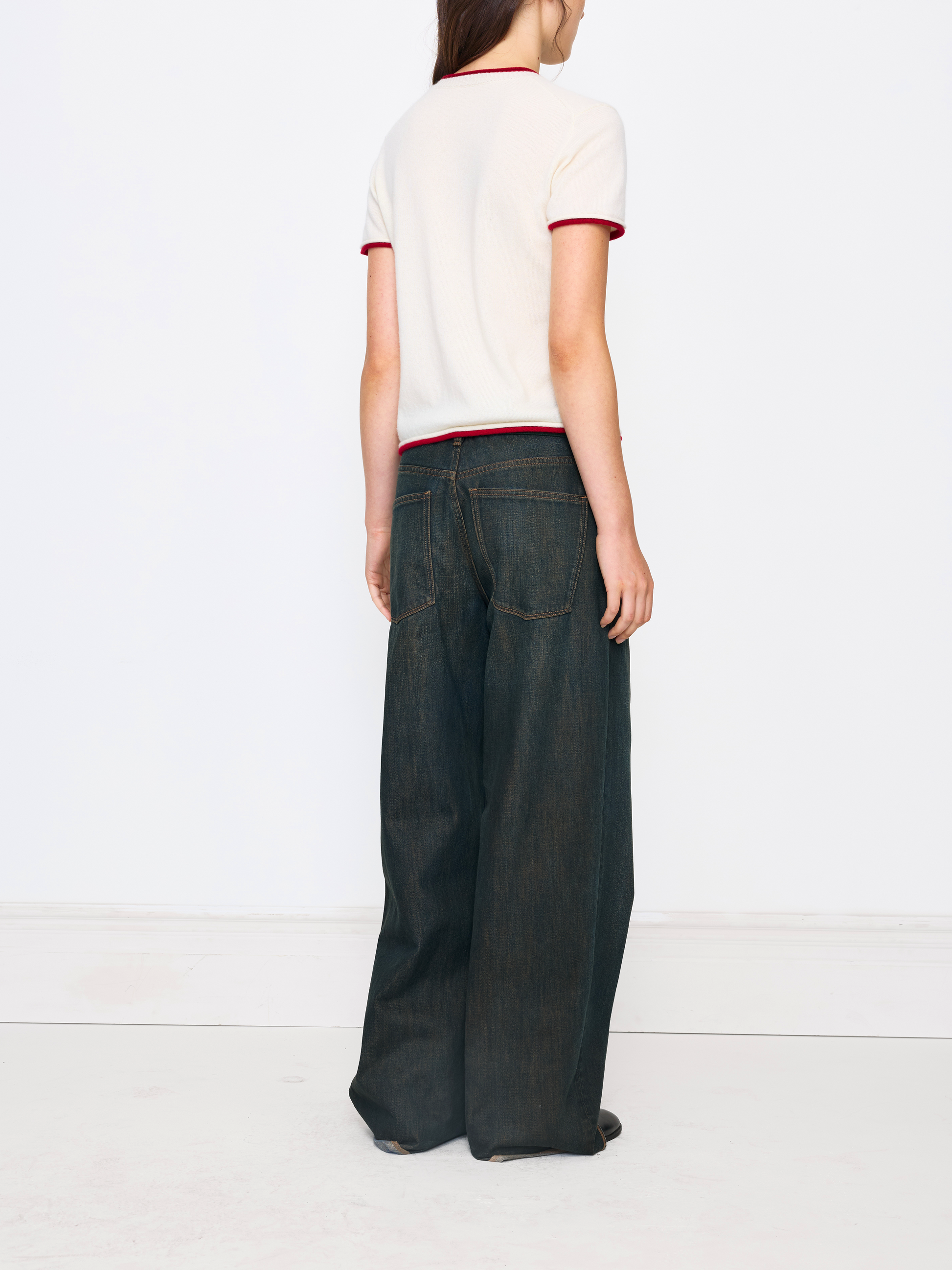 A model shown from the back wearing a cream short-sleeve knit top with red trim and dark wide-leg denim jeans, standing against a plain white studio background.