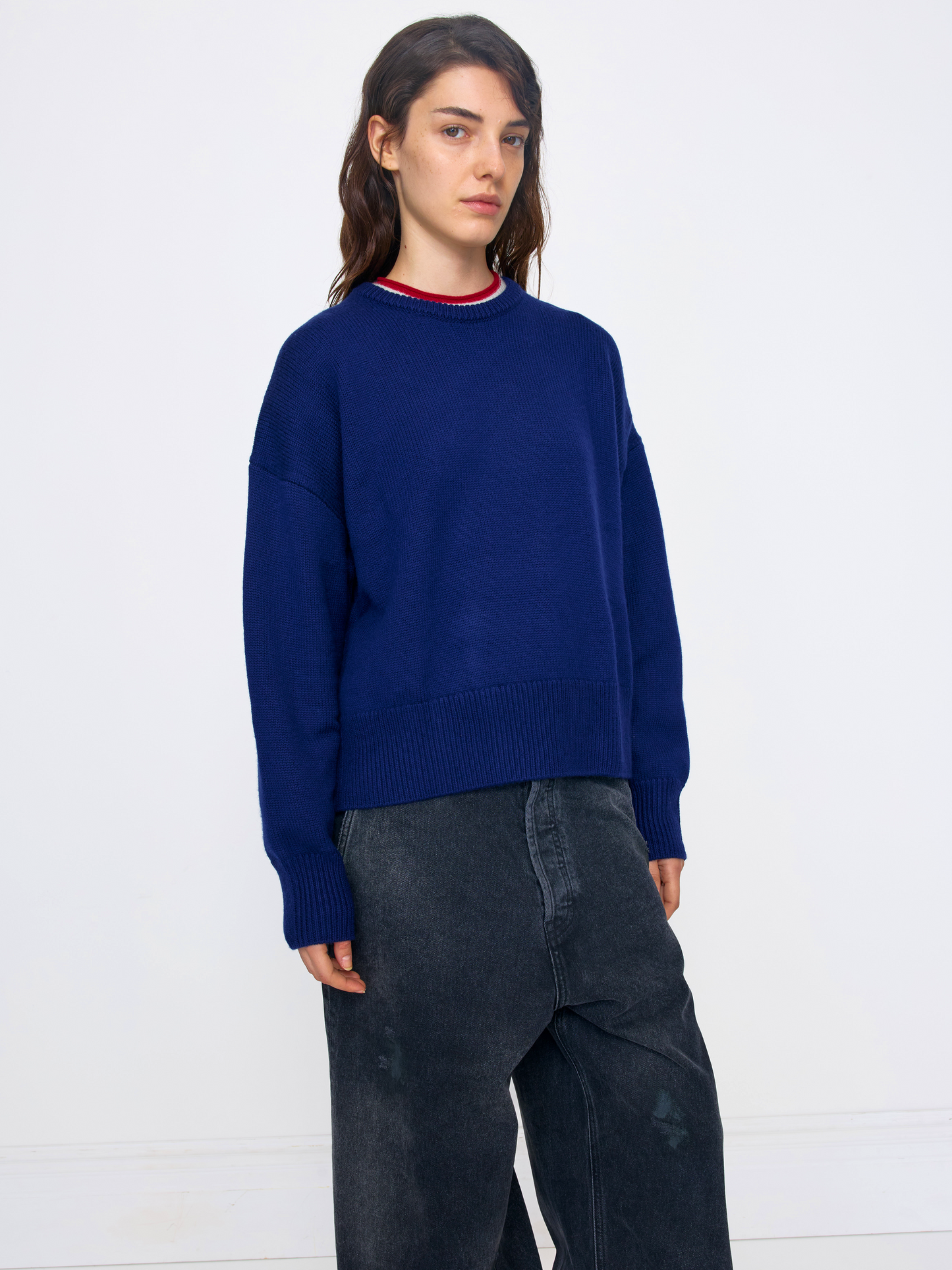 A woman modeling an oversized navy blue knit sweater with a red and white striped crew neck, paired with dark gray wide-leg jeans, against a plain white studio background.