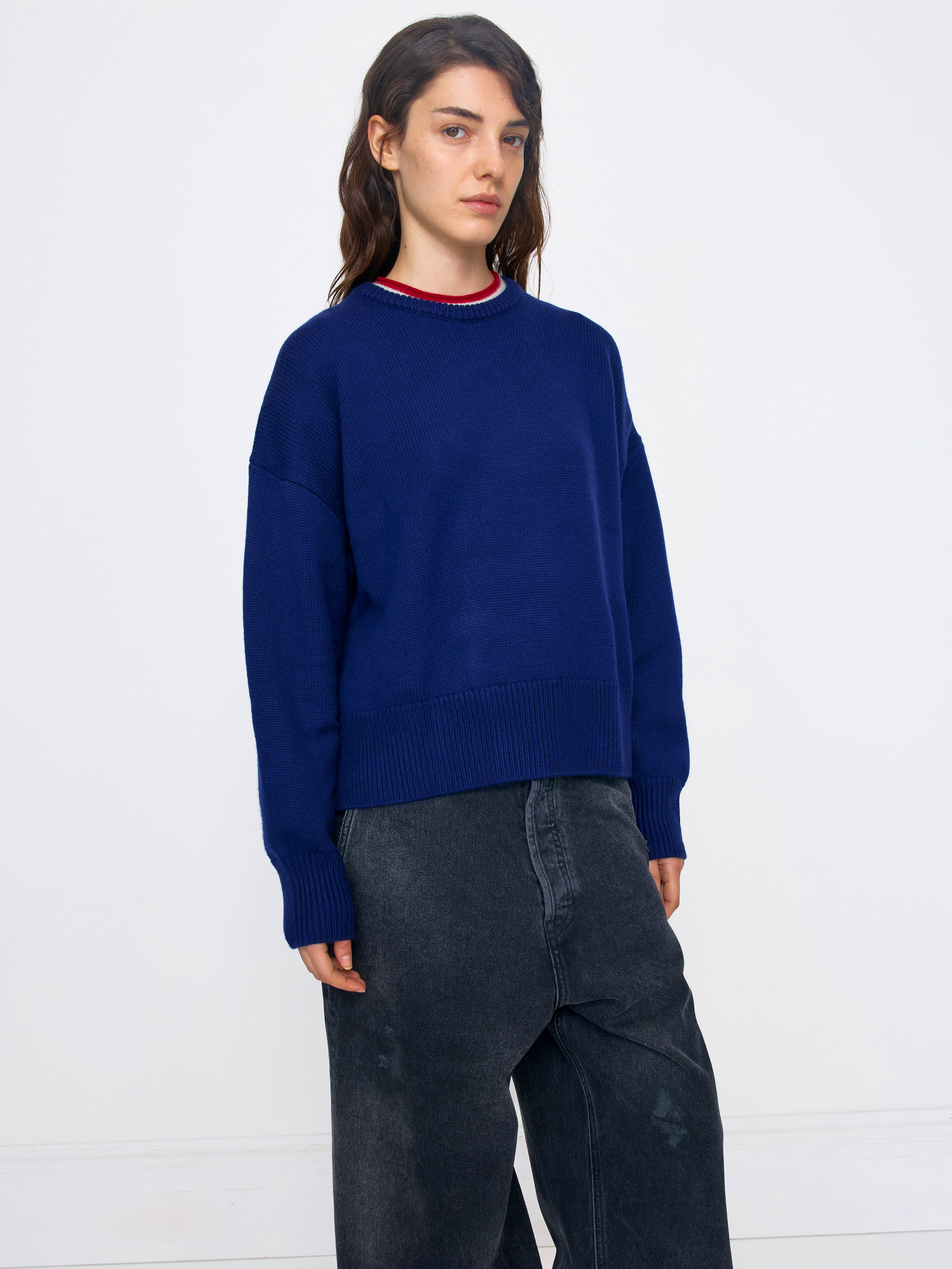 A woman modeling an oversized navy blue knit sweater with a red and white striped crew neck, paired with dark gray wide-leg jeans, against a plain white studio background.