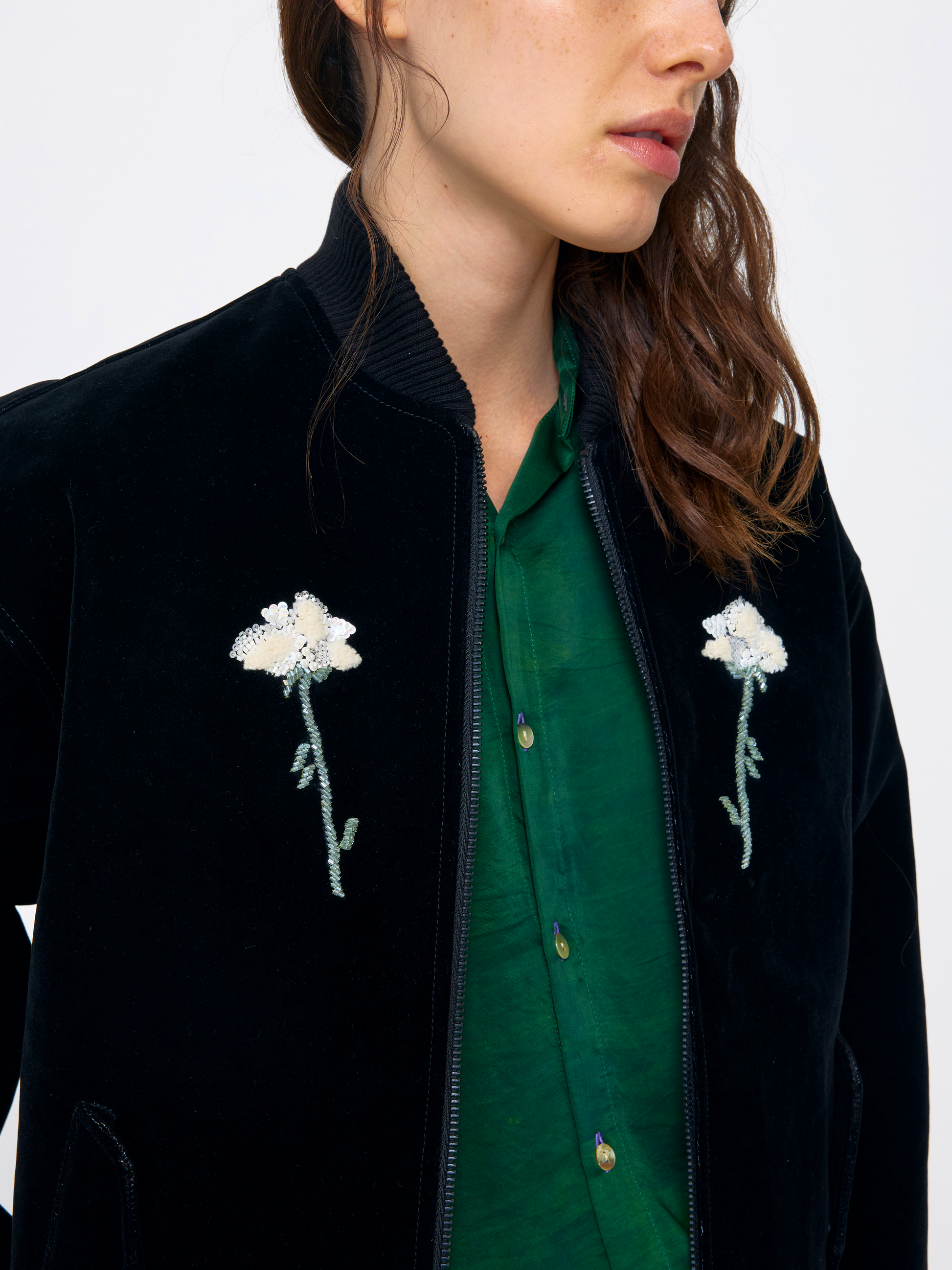 Close-up of a model wearing a black velvet bomber jacket with white floral embroidery on the chest, layered over a green button-up shirt.