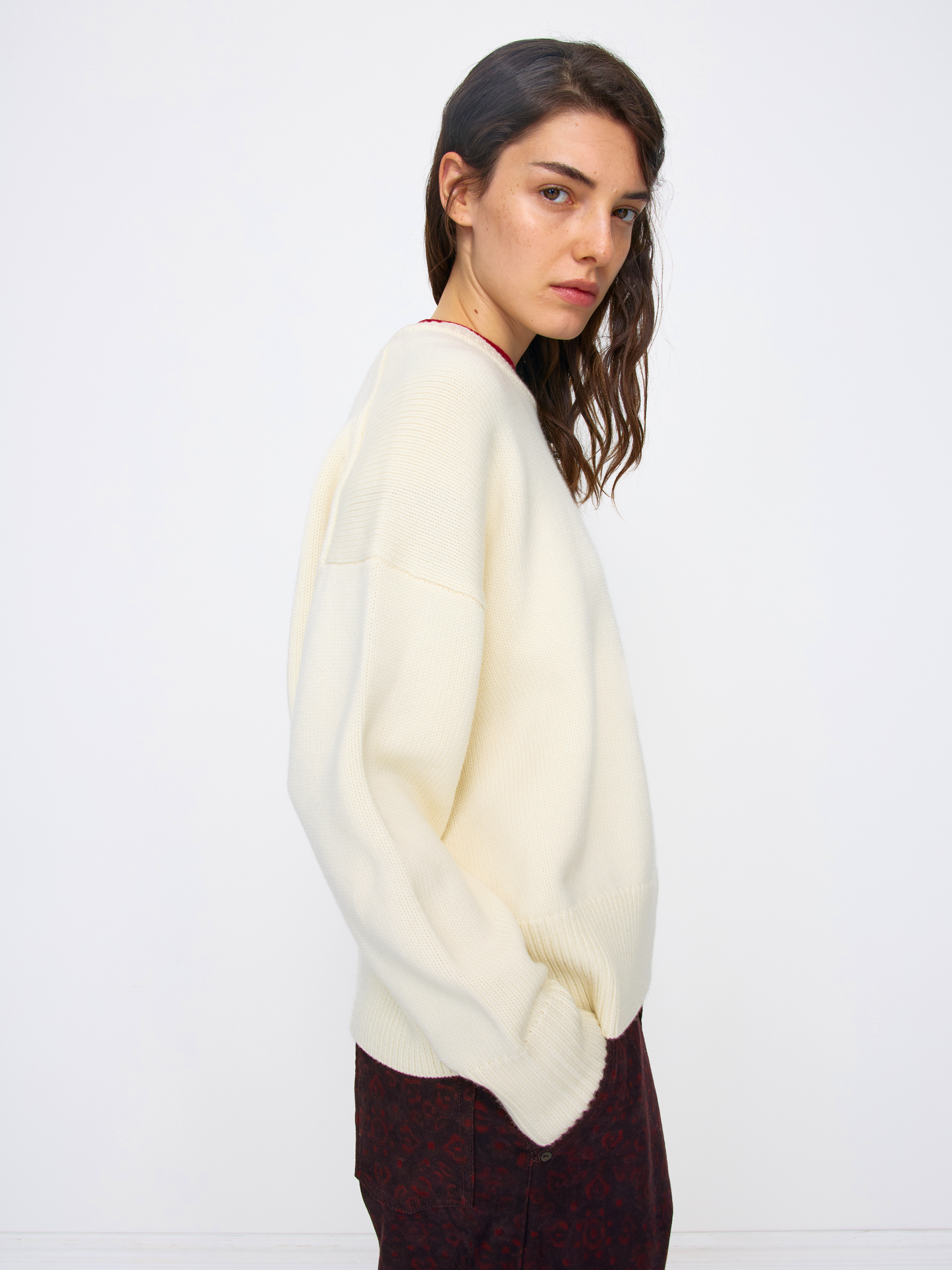 A woman wearing a cream-colored knit sweater and dark patterned trousers, photographed against a plain white studio background.