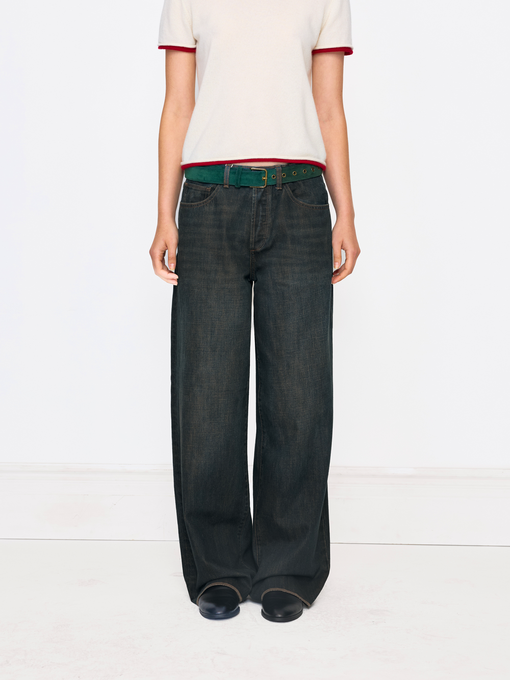 A model is shown from the chest down wearing dark wash wide-leg jeans, a green belt, a cream ringer t-shirt with red trim, and black shoes, set against a plain white studio background.
