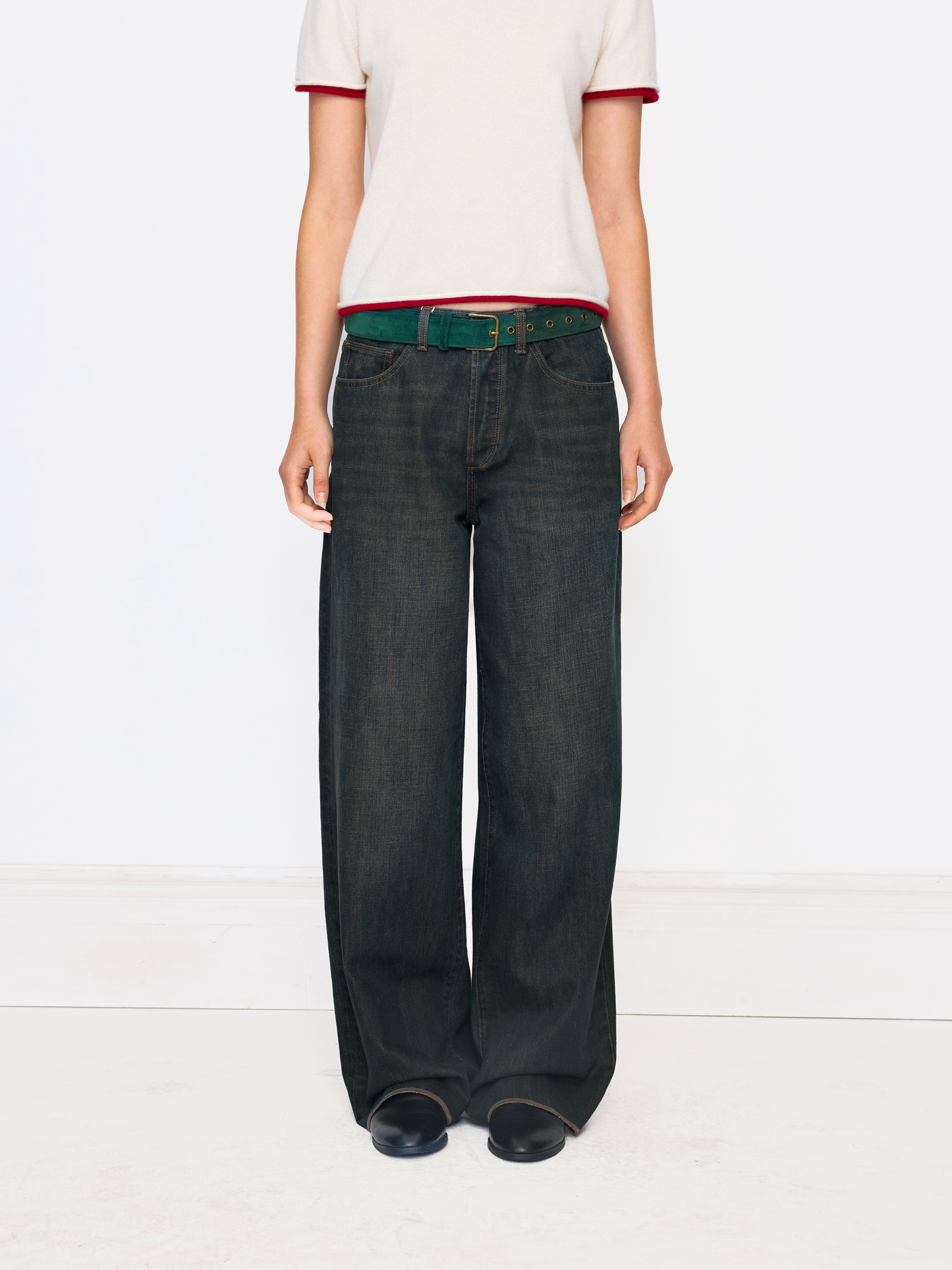 A model is shown from the chest down wearing dark wash wide-leg jeans, a green belt, a cream ringer t-shirt with red trim, and black shoes, set against a plain white studio background.