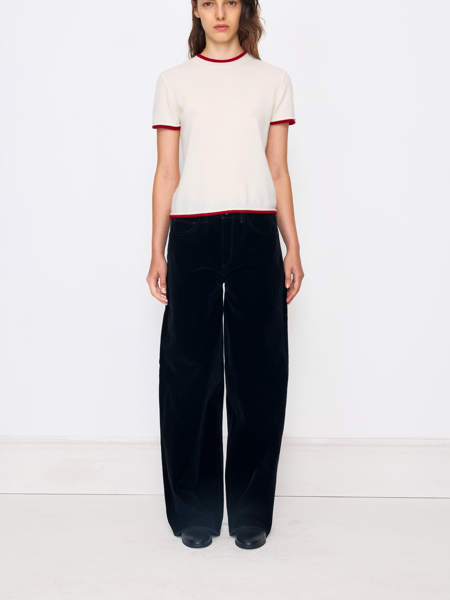 A model stands against a plain white studio wall wearing a cream short-sleeve knit top with red trim and black wide-leg velvet pants.