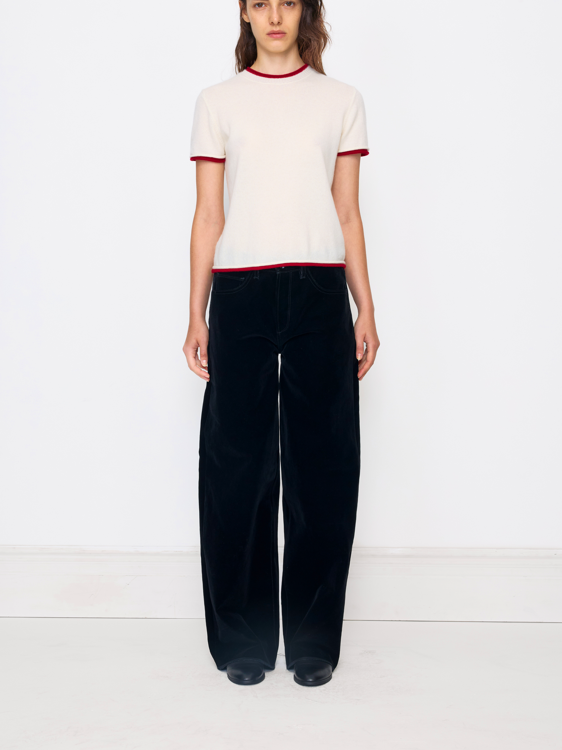 A model stands against a plain white studio wall wearing a cream short-sleeve knit top with red trim and black wide-leg velvet pants.