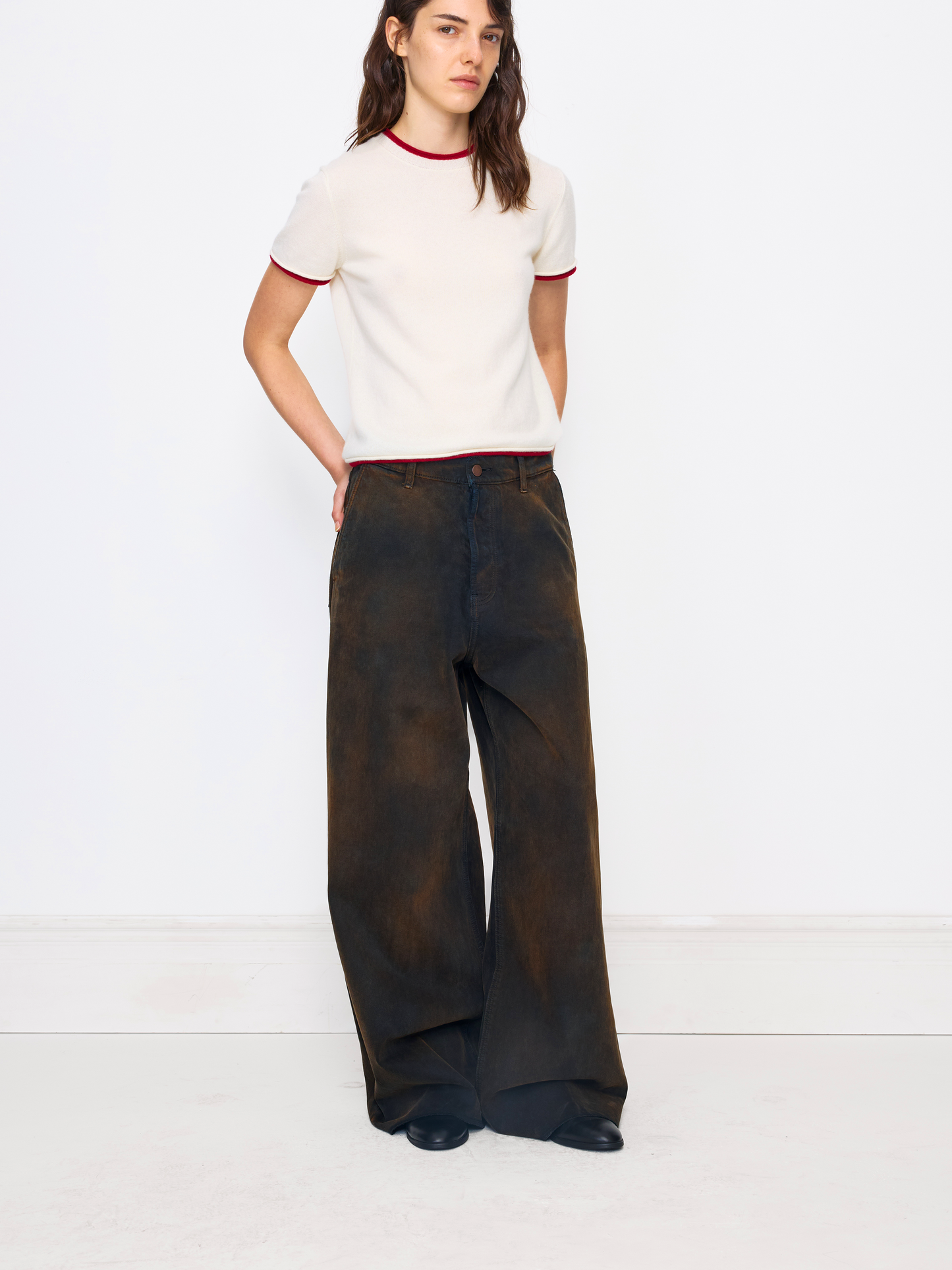 A model stands against a white studio wall wearing a cream short-sleeve knit top with red trim and very wide-leg dark brown pants.