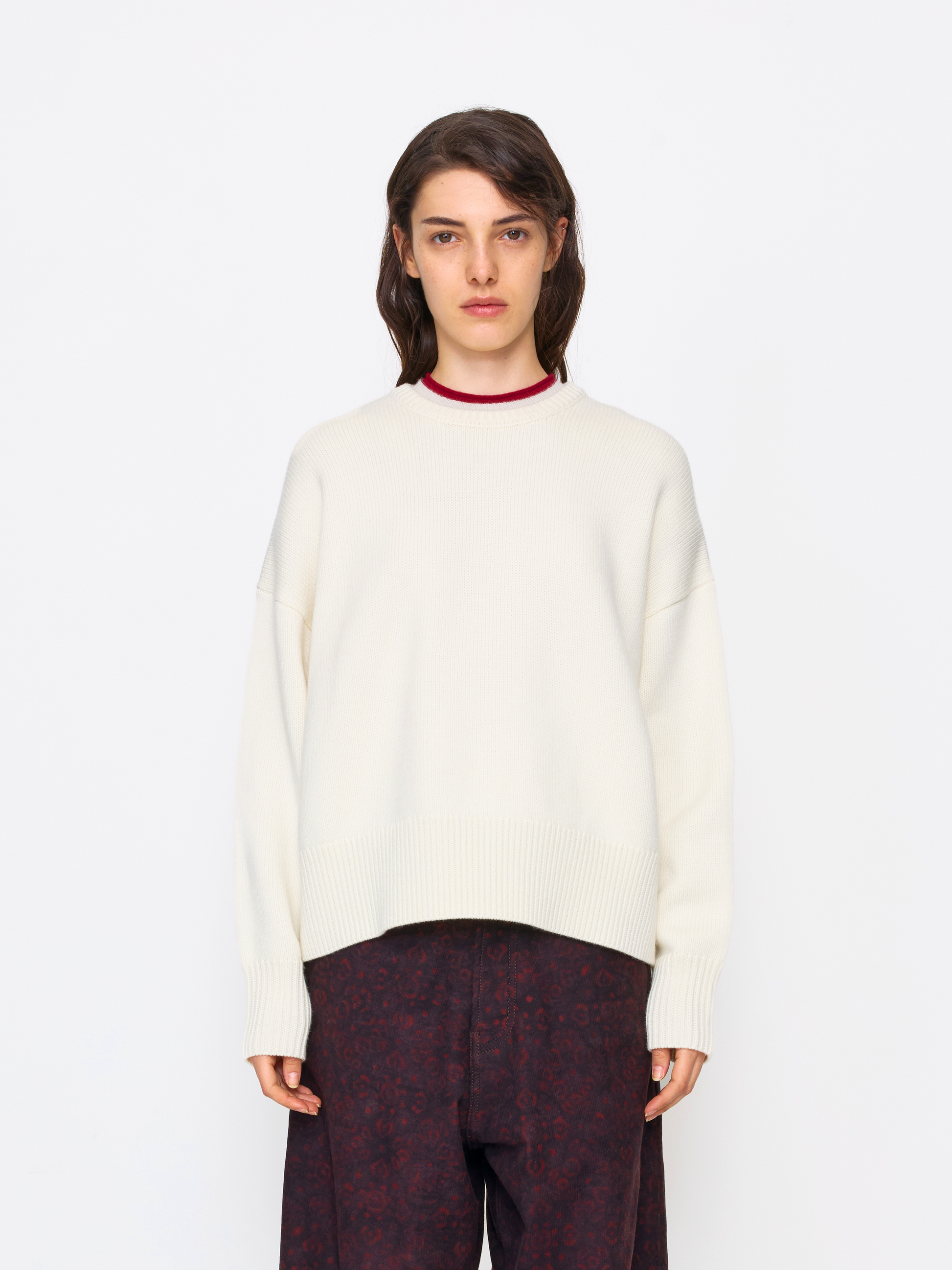 A model wearing an oversized cream-colored knit sweater with a contrasting dark red crew neck, paired with dark patterned trousers, shot against a solid white studio background.