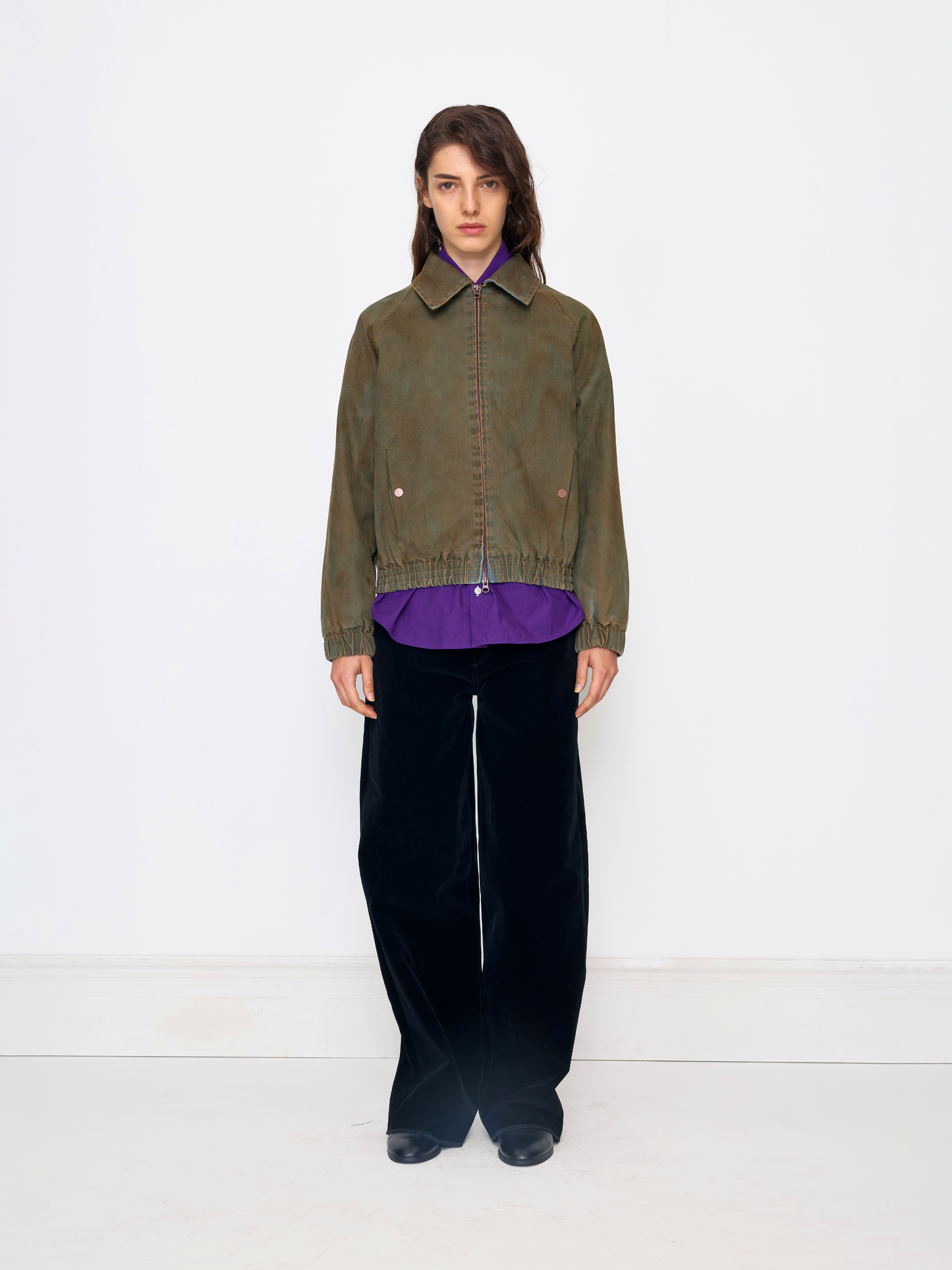 A model stands against a plain white studio background wearing an olive green cropped jacket over a purple shirt, paired with black wide-leg trousers and black shoes.