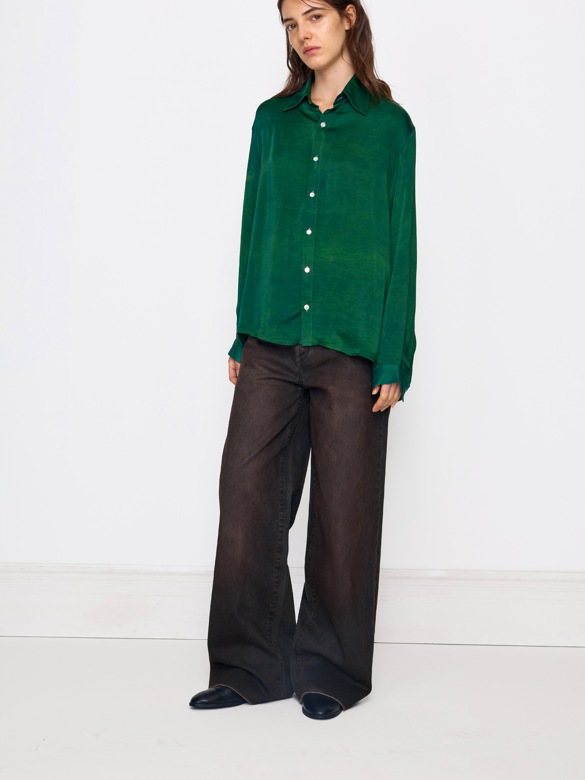 A woman modeling a long-sleeve green button-up shirt with white buttons, paired with dark brown wide-leg trousers and black shoes, standing against a plain white studio background.