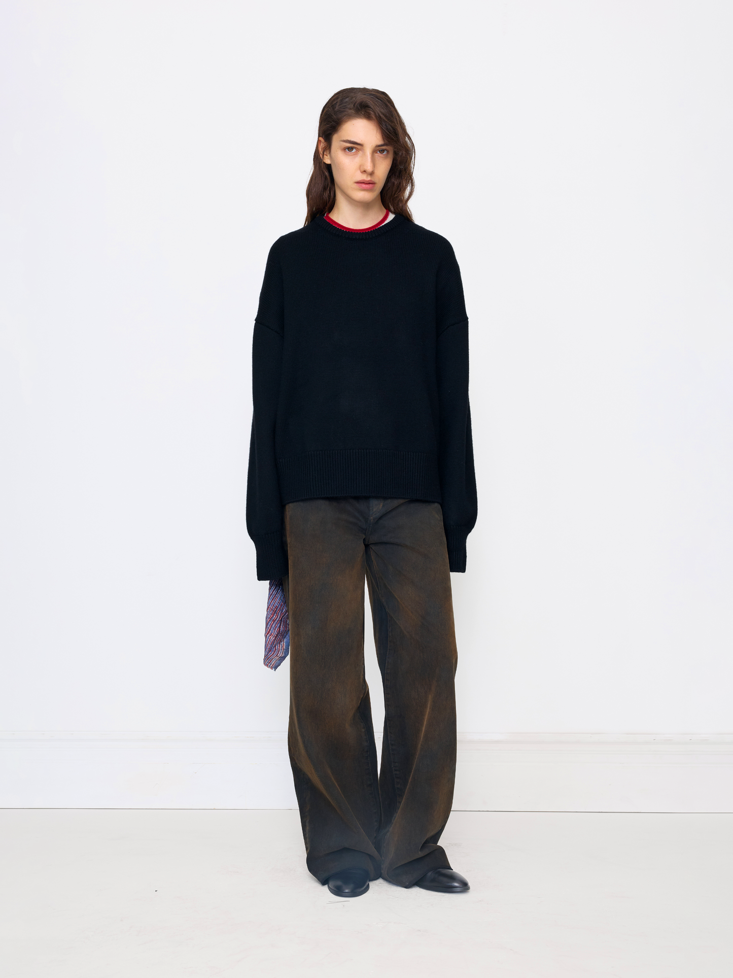 A model stands against a plain white studio background wearing an oversized black knit sweater with a red crew neck, wide-leg brown trousers, and black shoes.