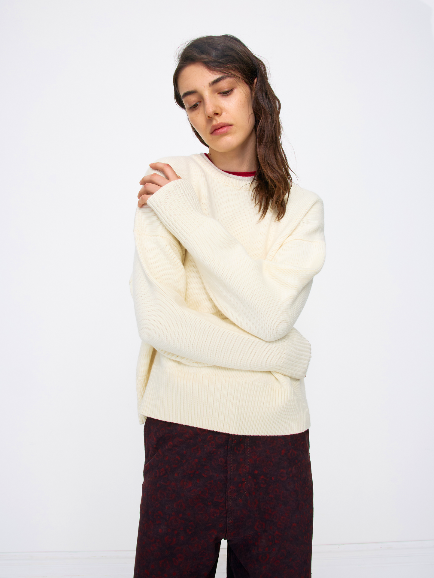 A woman wearing an oversized cream-colored ribbed knit sweater and dark patterned trousers, standing against a plain white studio background.