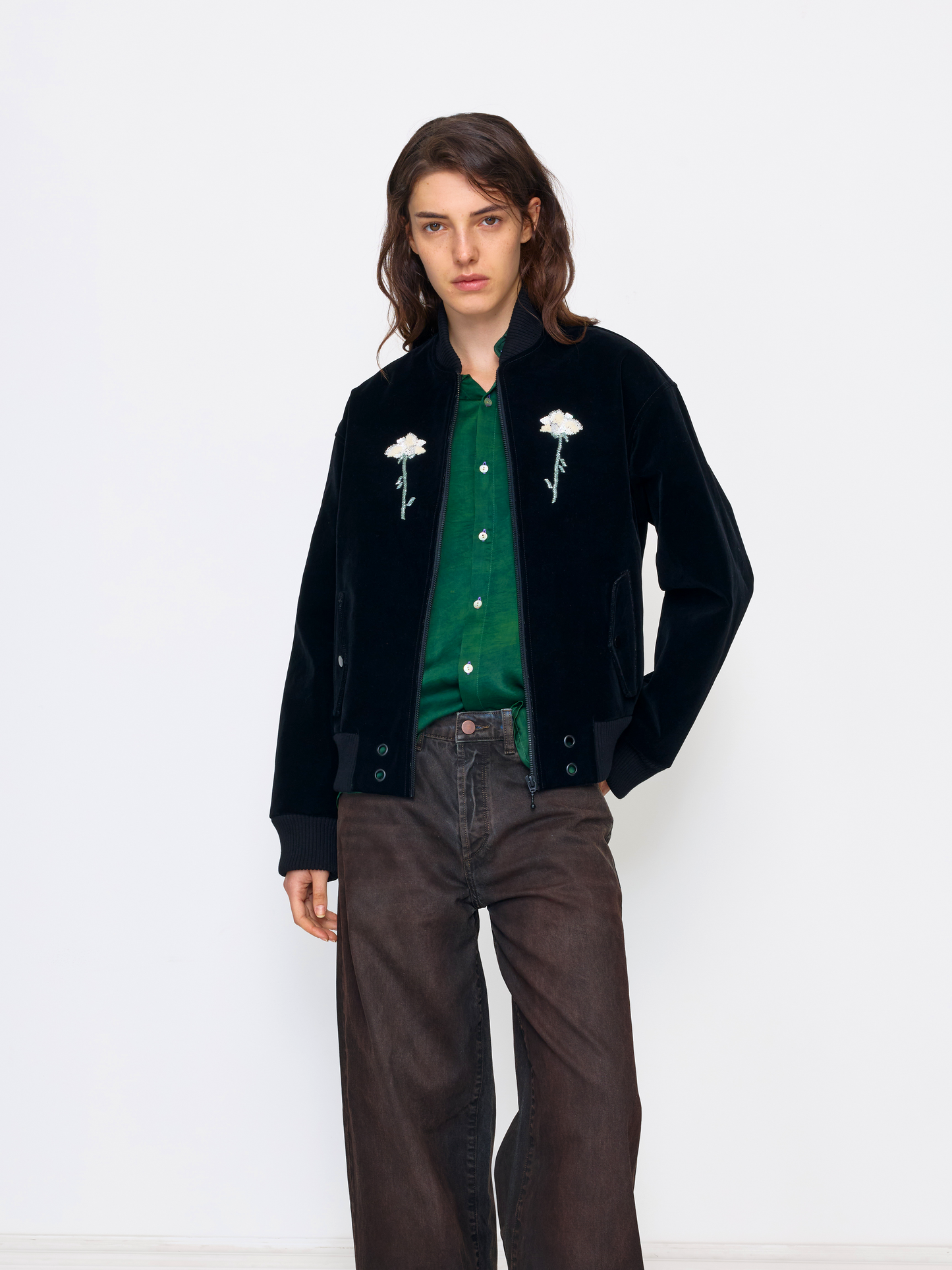 A model stands against a plain white studio background wearing a black velvet jacket with white embroidered flowers, a green knit top, and dark brown wide-leg jeans.