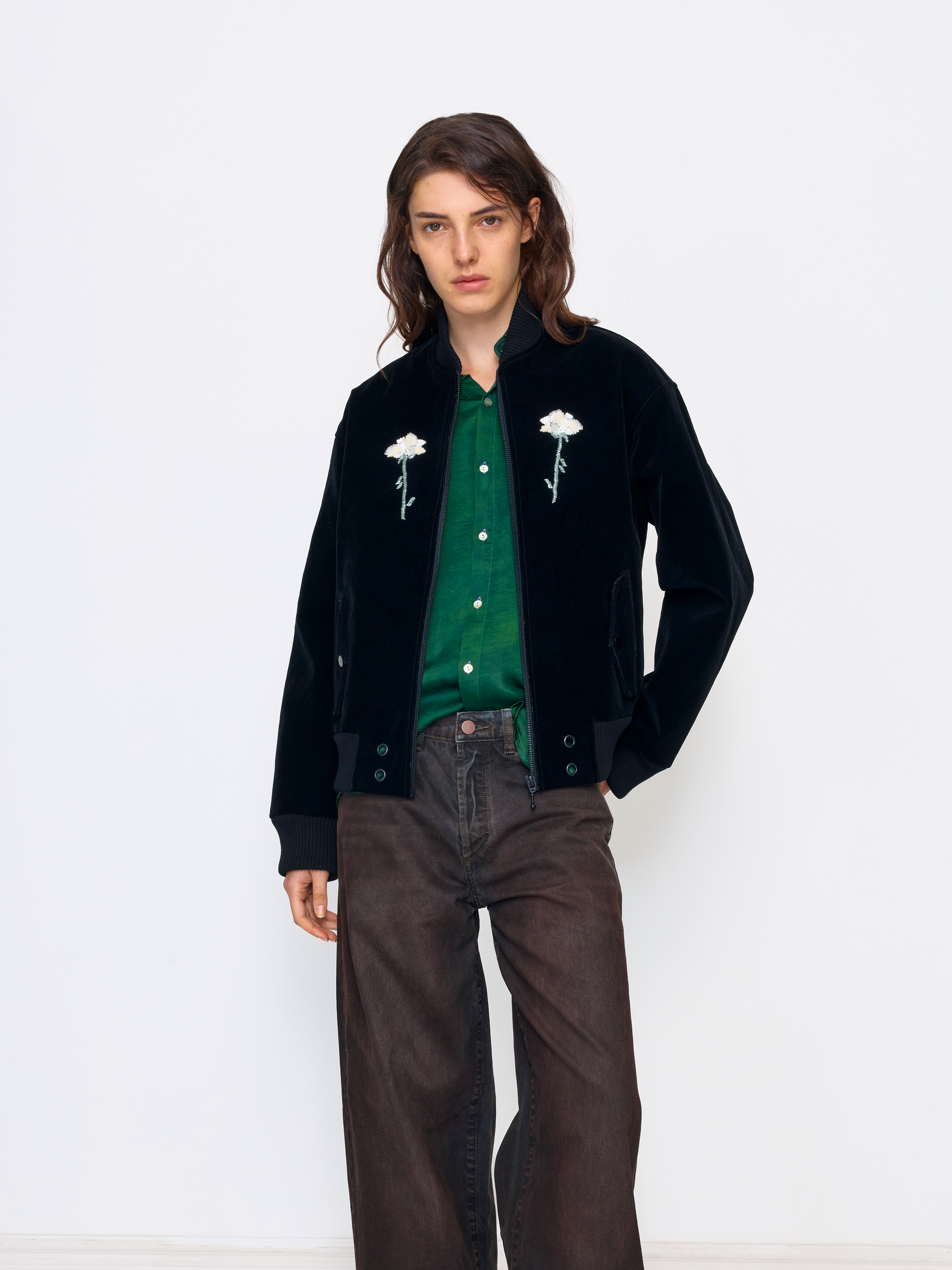 A model stands against a plain white studio background wearing a black velvet jacket with white embroidered flowers, a green knit top, and dark brown wide-leg jeans.