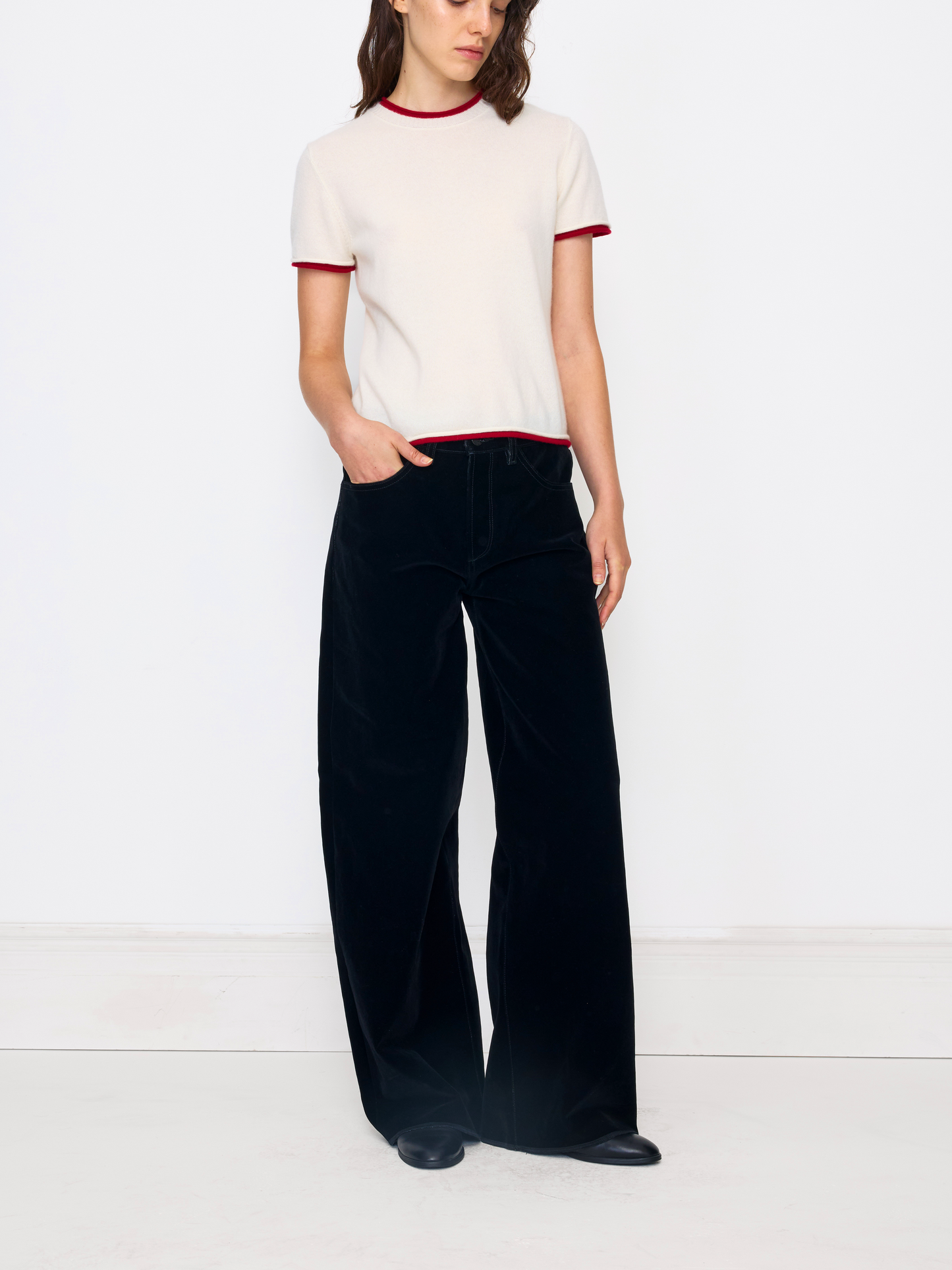 A model wearing a cream short-sleeve knit top with red trim and black wide-leg trousers, standing against a plain white studio background.