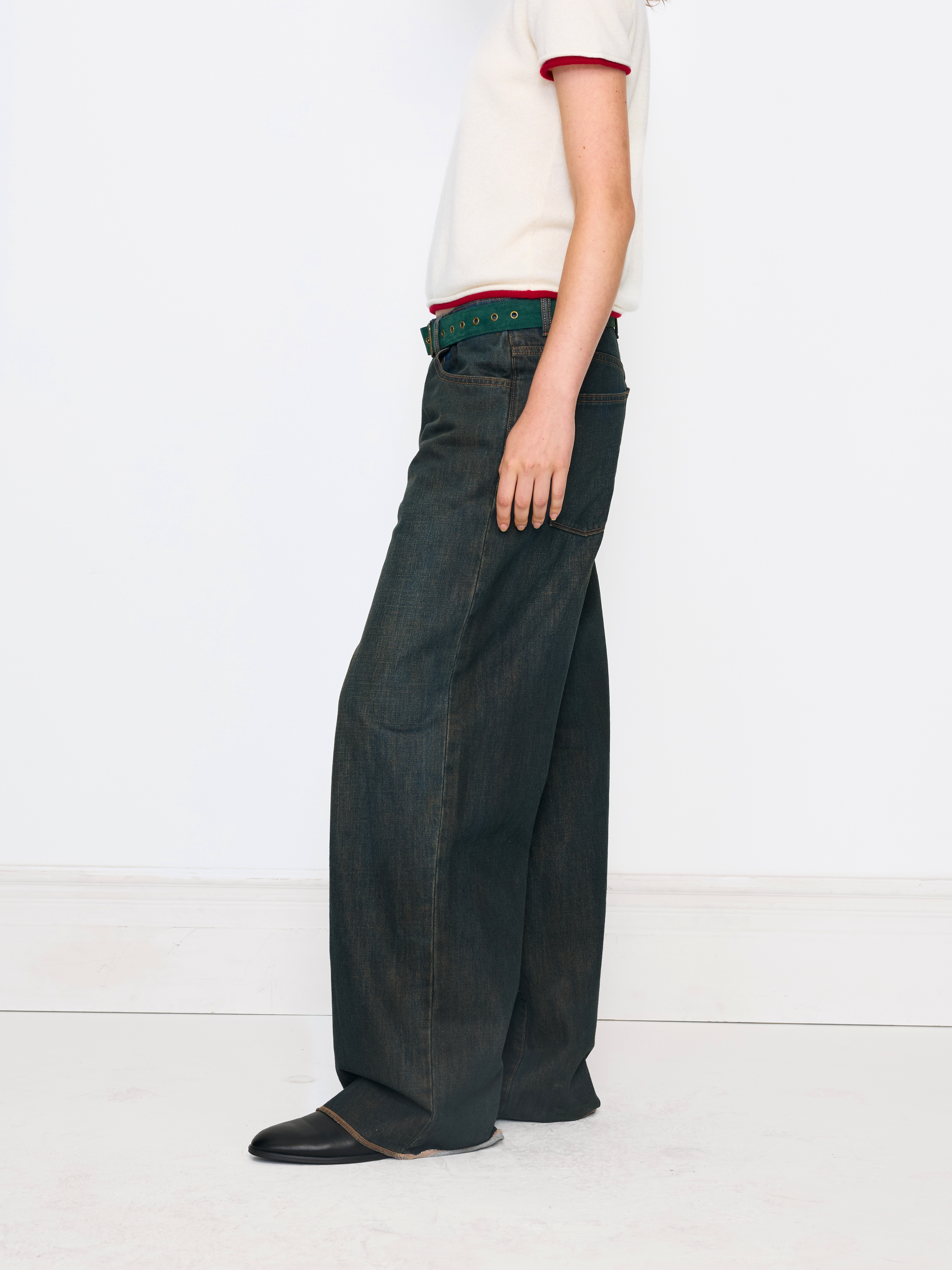 A side-profile view of a model wearing dark indigo wide-leg jeans, a green belt, and a cream short-sleeve top against a clean white studio background.