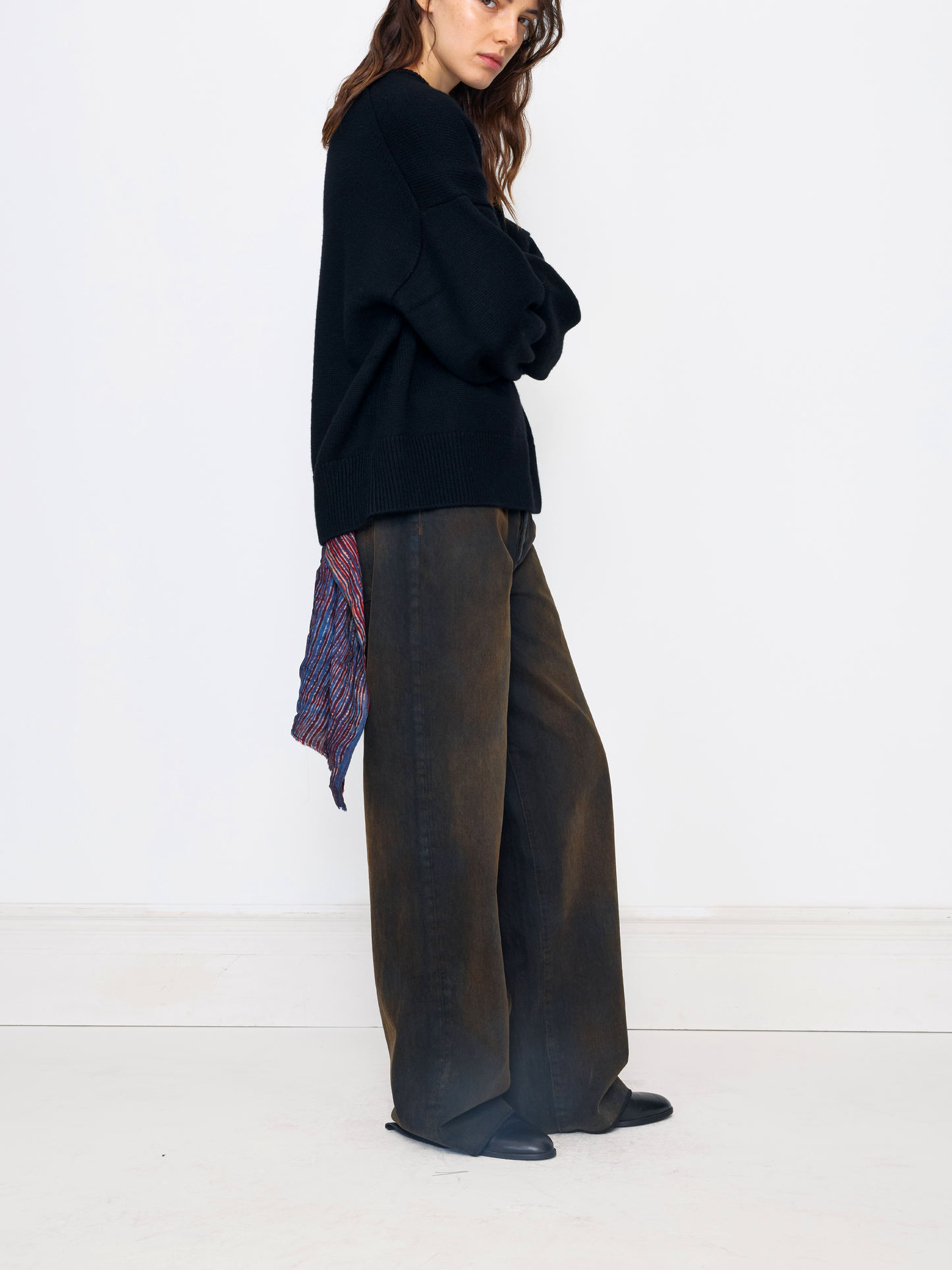 A model is shown from the side in a studio setting, wearing a black knit sweater, wide-leg dark brown trousers, and black shoes. A patterned scarf is draped over her shoulder.