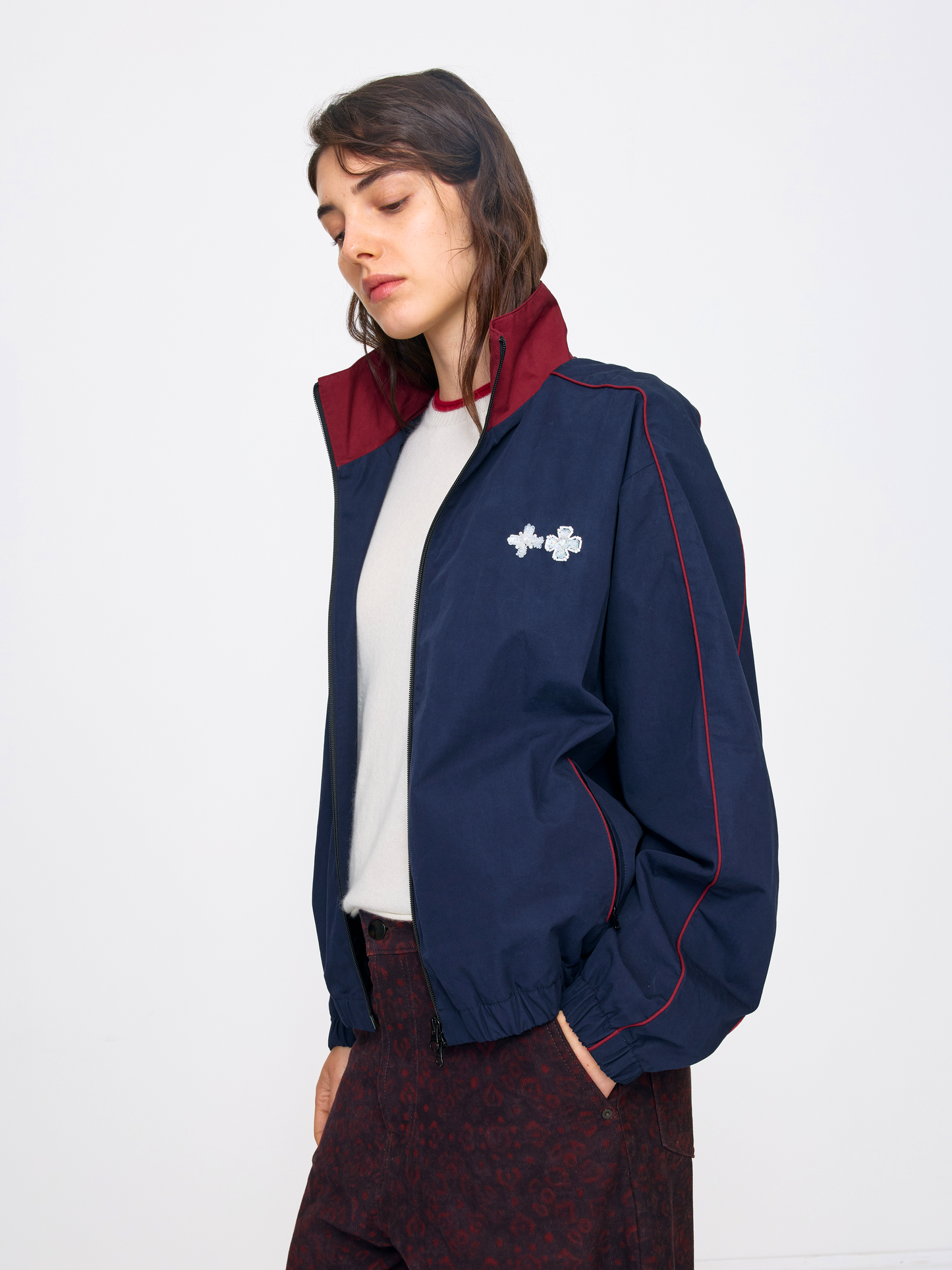 A woman wearing a navy blue windbreaker jacket with a burgundy collar and red piping, styled over a white t-shirt and dark patterned trousers, photographed against a plain white studio background.