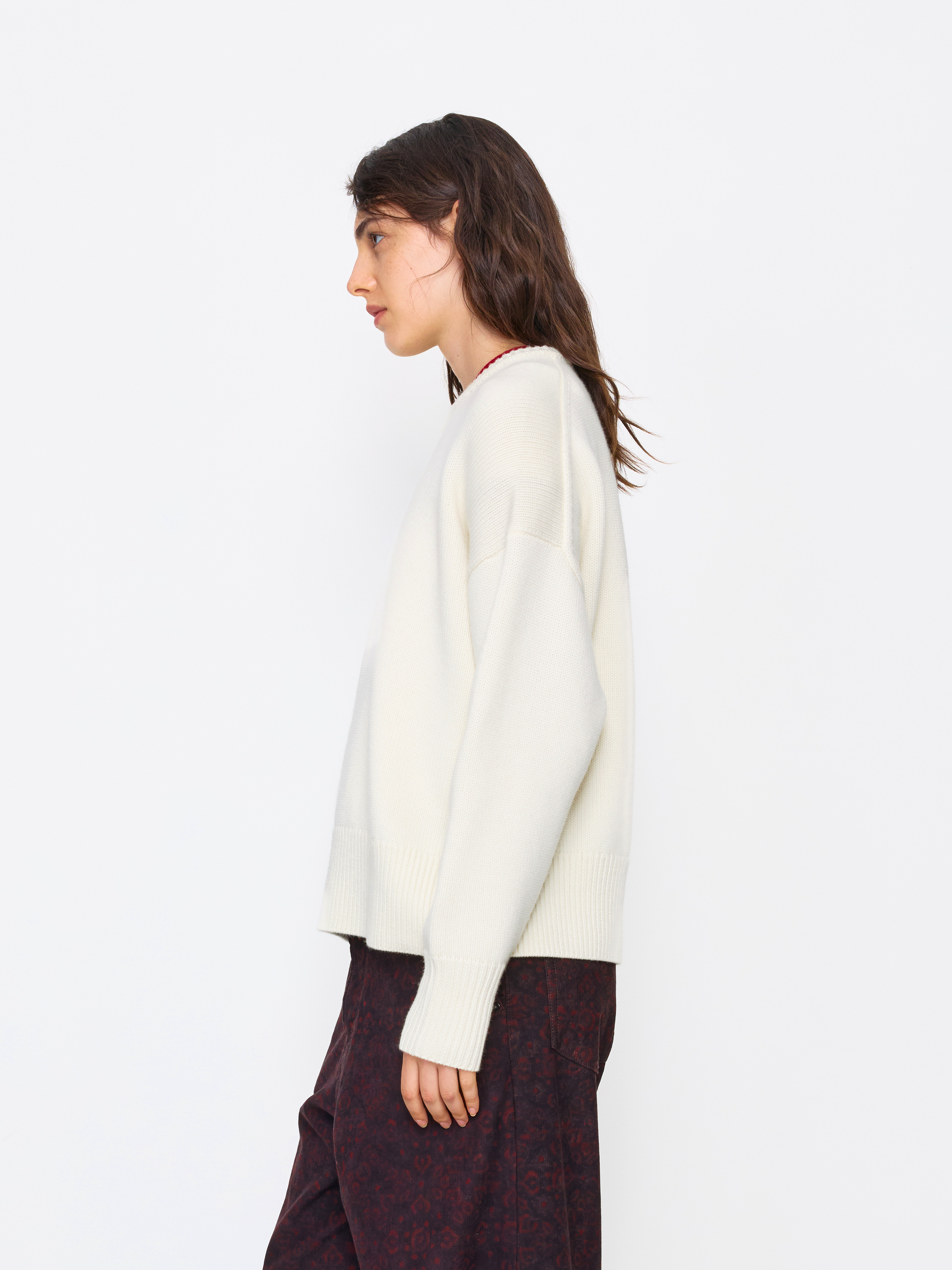A woman shown in side profile wearing a cream knit sweater and dark patterned pants against a plain light gray studio background.