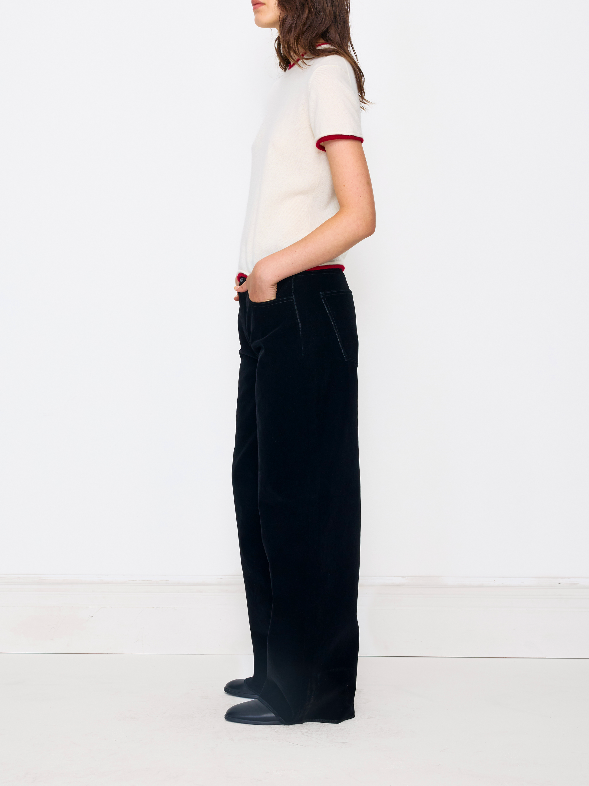 A model shown from the side profile wearing black wide-leg trousers, a cream short-sleeve knit top with red trim, and dark loafers, set against a clean white studio background.
