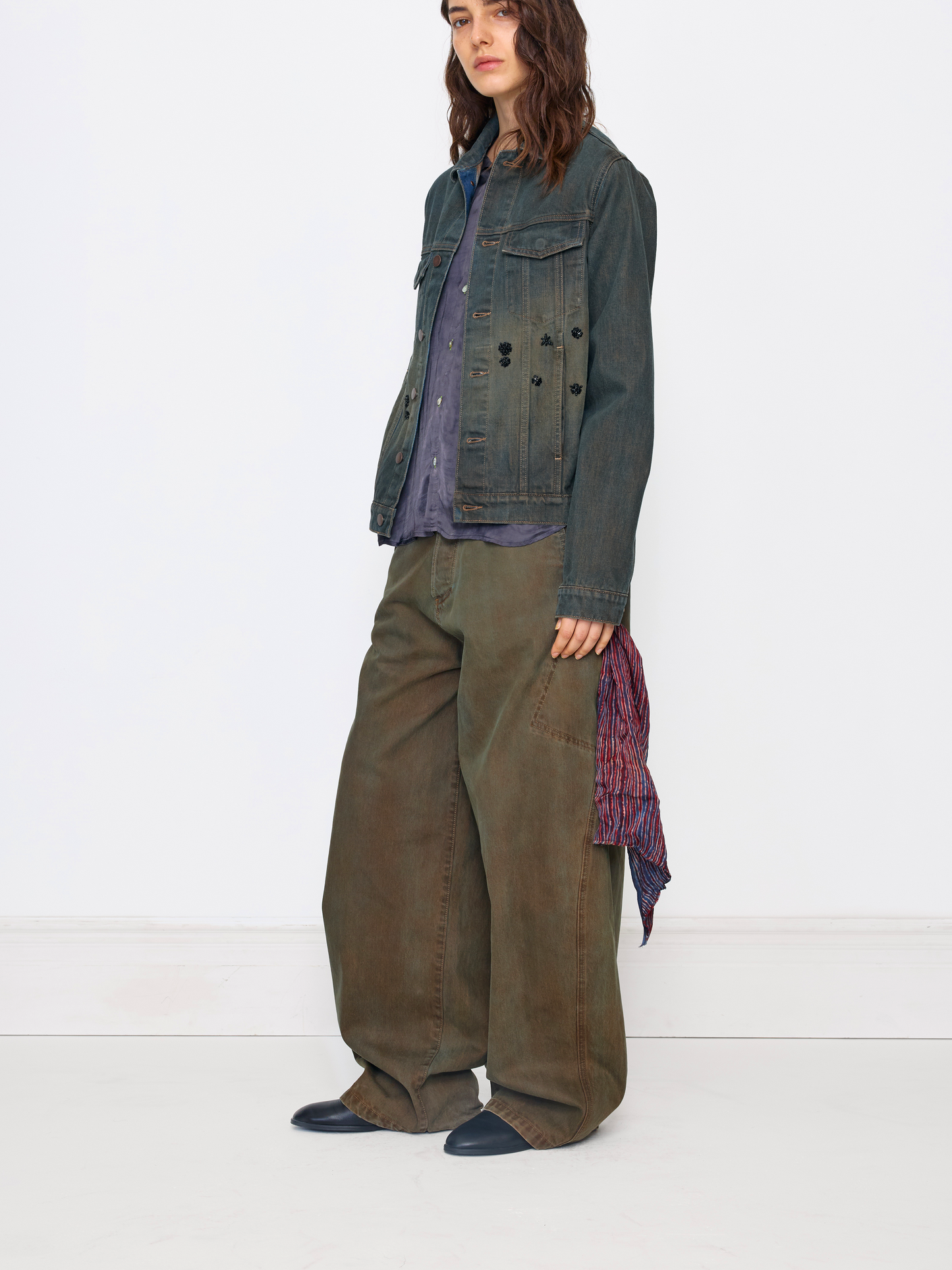 A model stands against a white studio background wearing a dark washed denim jacket over a purple shirt, wide-leg olive pants, and dark slip-on shoes, with a patterned scarf draped from one hand.