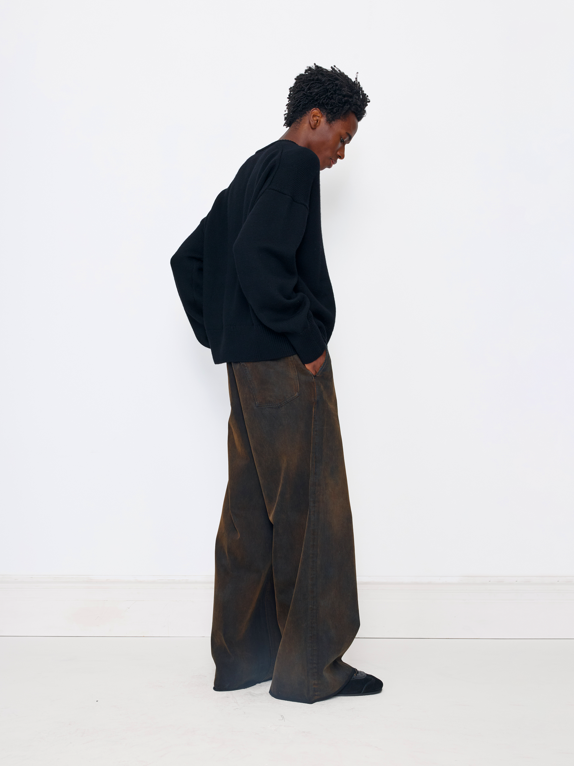 A model wearing a black oversized long-sleeve top and wide-leg brown pants with a faded, distressed texture, standing in profile against a plain white studio wall.