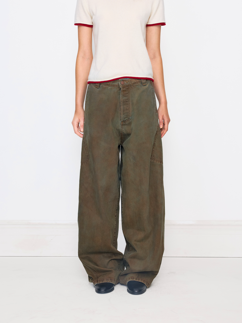 A model is shown from the chest down wearing wide-leg, relaxed-fit brown pants with a cream t-shirt featuring red trim, standing against a plain white studio background.