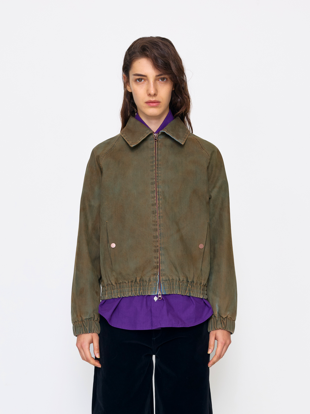 A model wearing an olive green zip-up jacket with a collar and elastic hem, layered over a purple shirt and paired with black pants, shot against a plain white studio background.