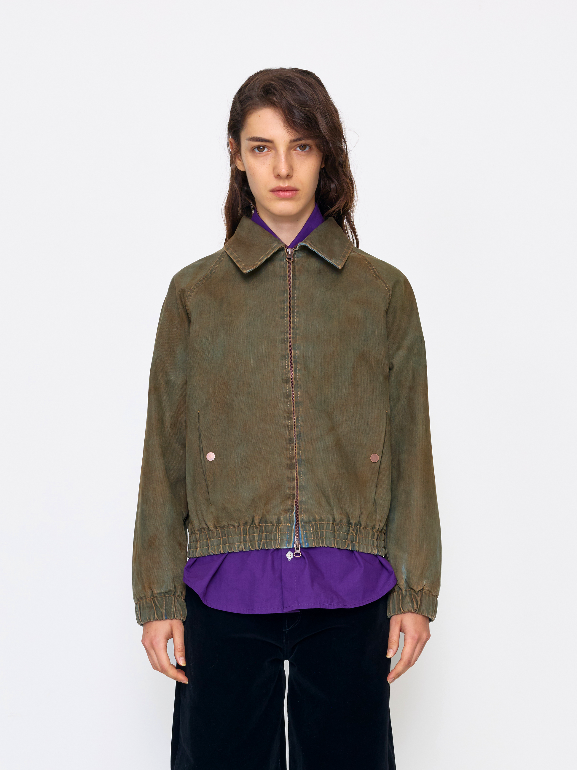 A model wearing an olive green zip-up jacket with a collar and elastic hem, layered over a purple shirt and paired with black pants, shot against a plain white studio background.