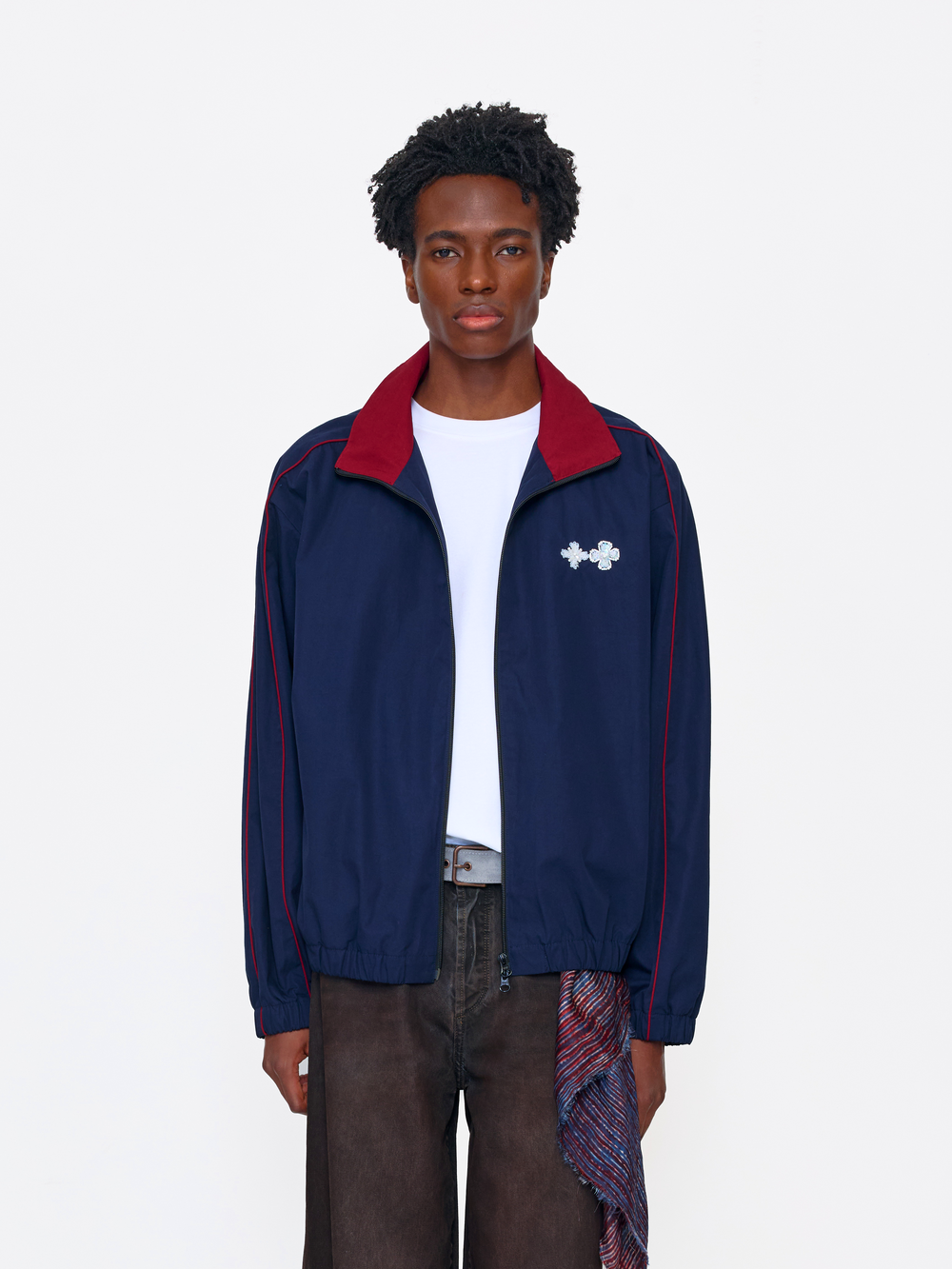A model wearing a navy blue track jacket with a red collar and red piping, featuring a white Under Armour logo on the chest, styled over a white t-shirt and dark brown pants.