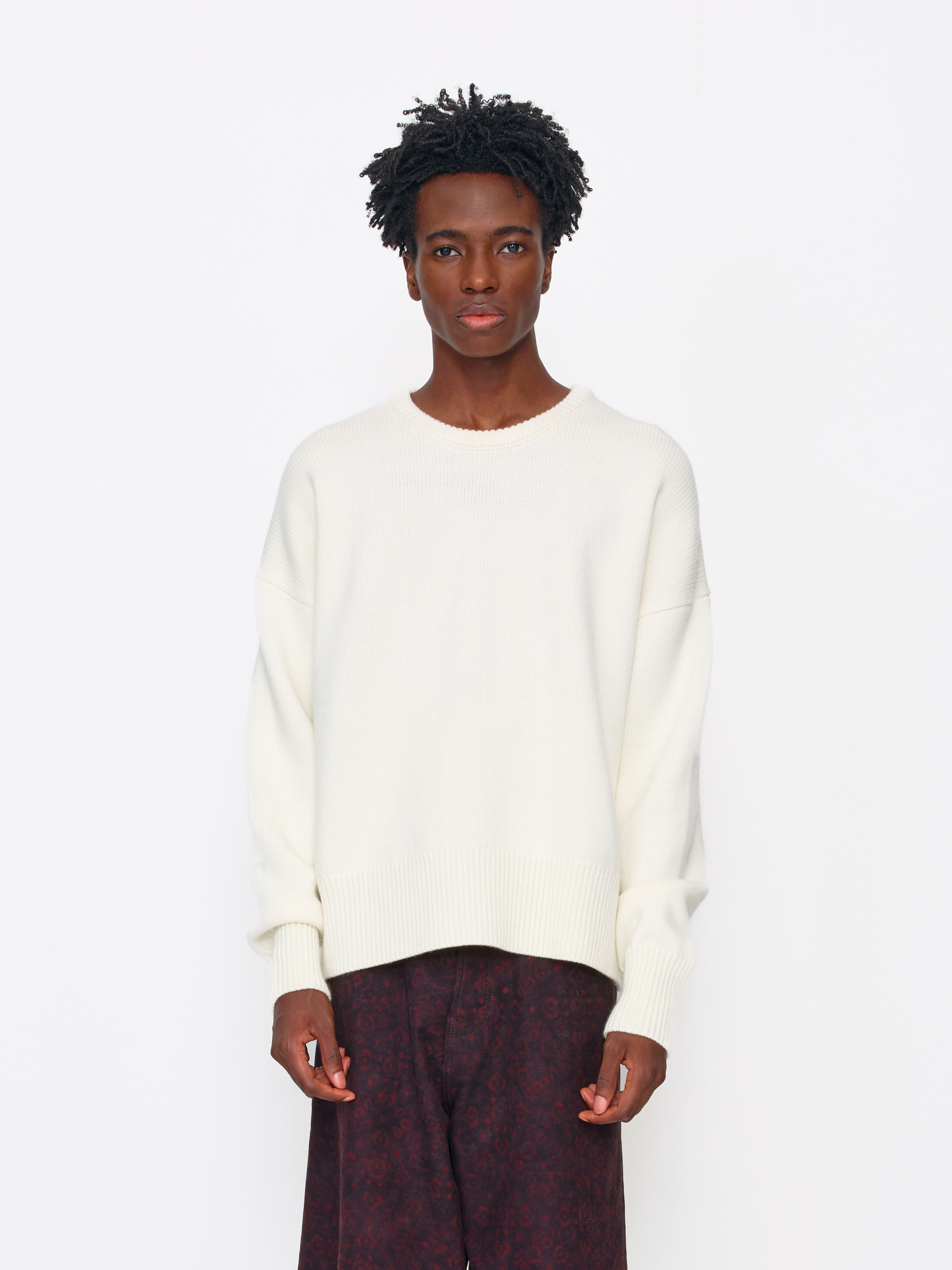 A model wearing an oversized cream-colored knit crewneck sweater and dark patterned trousers, standing against a plain white studio background.