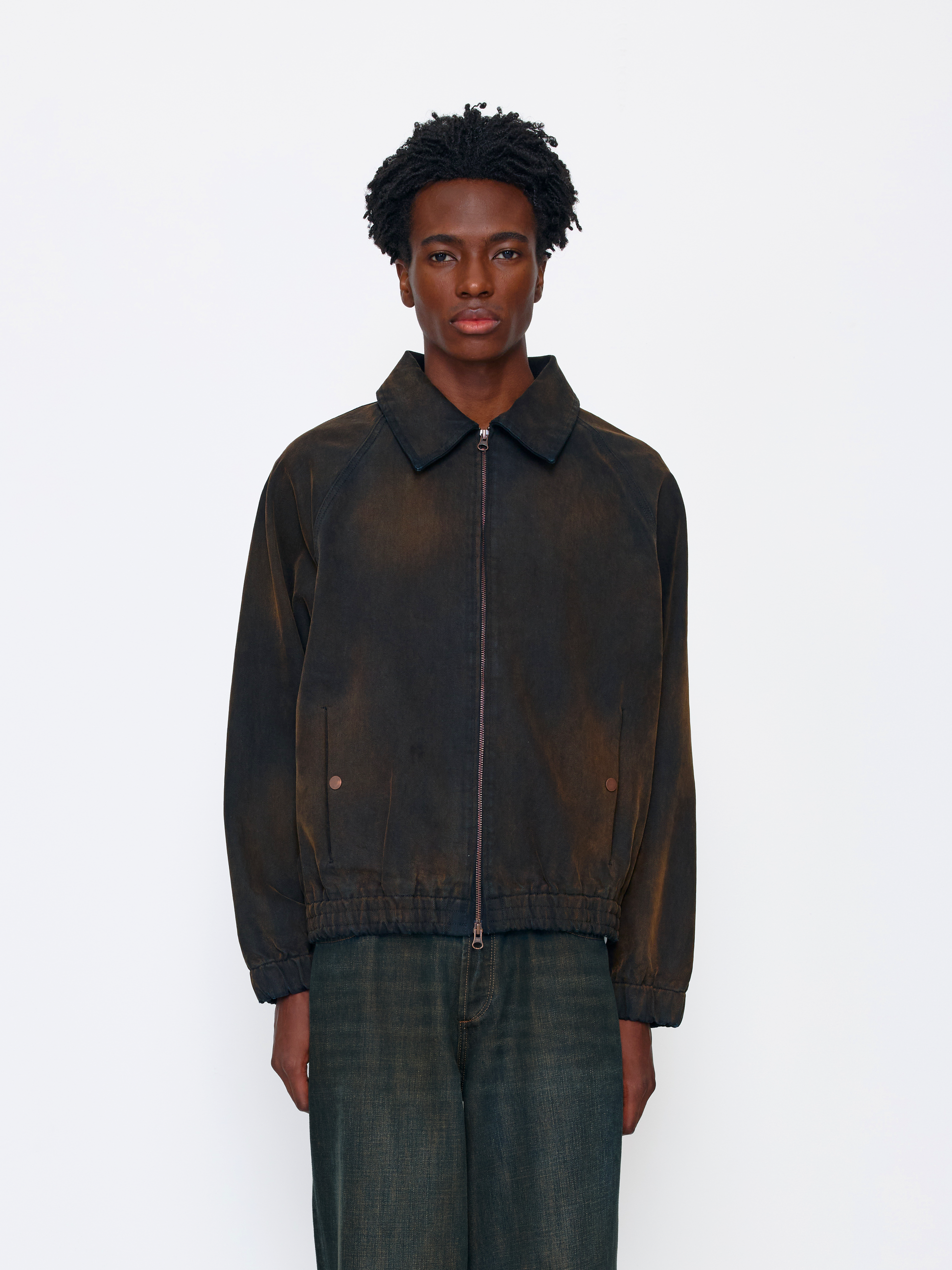 A model wearing a dark brown and black acid-washed zip-up jacket with a pointed collar, paired with dark green jeans, against a plain white studio background.