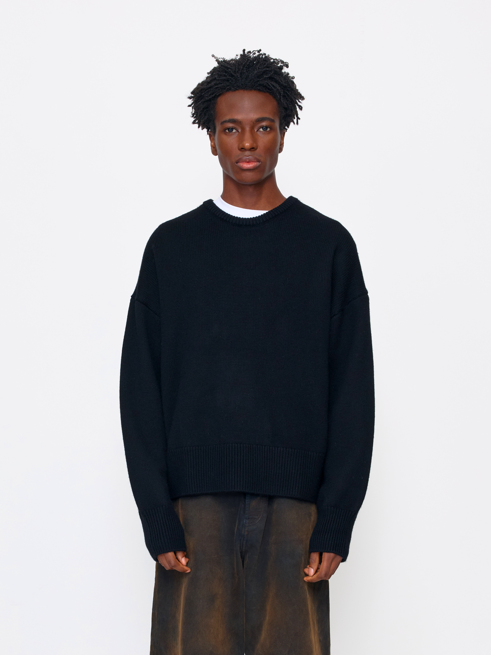 A model wearing a black knit crewneck sweater over a white undershirt and brown trousers, standing against a plain white studio background.