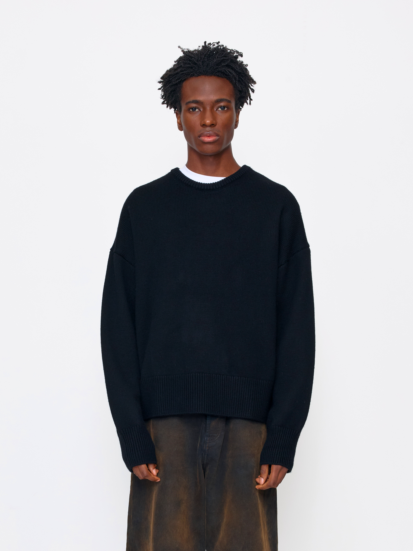 A model wearing a black knit crewneck sweater over a white undershirt and brown trousers, standing against a plain white studio background.