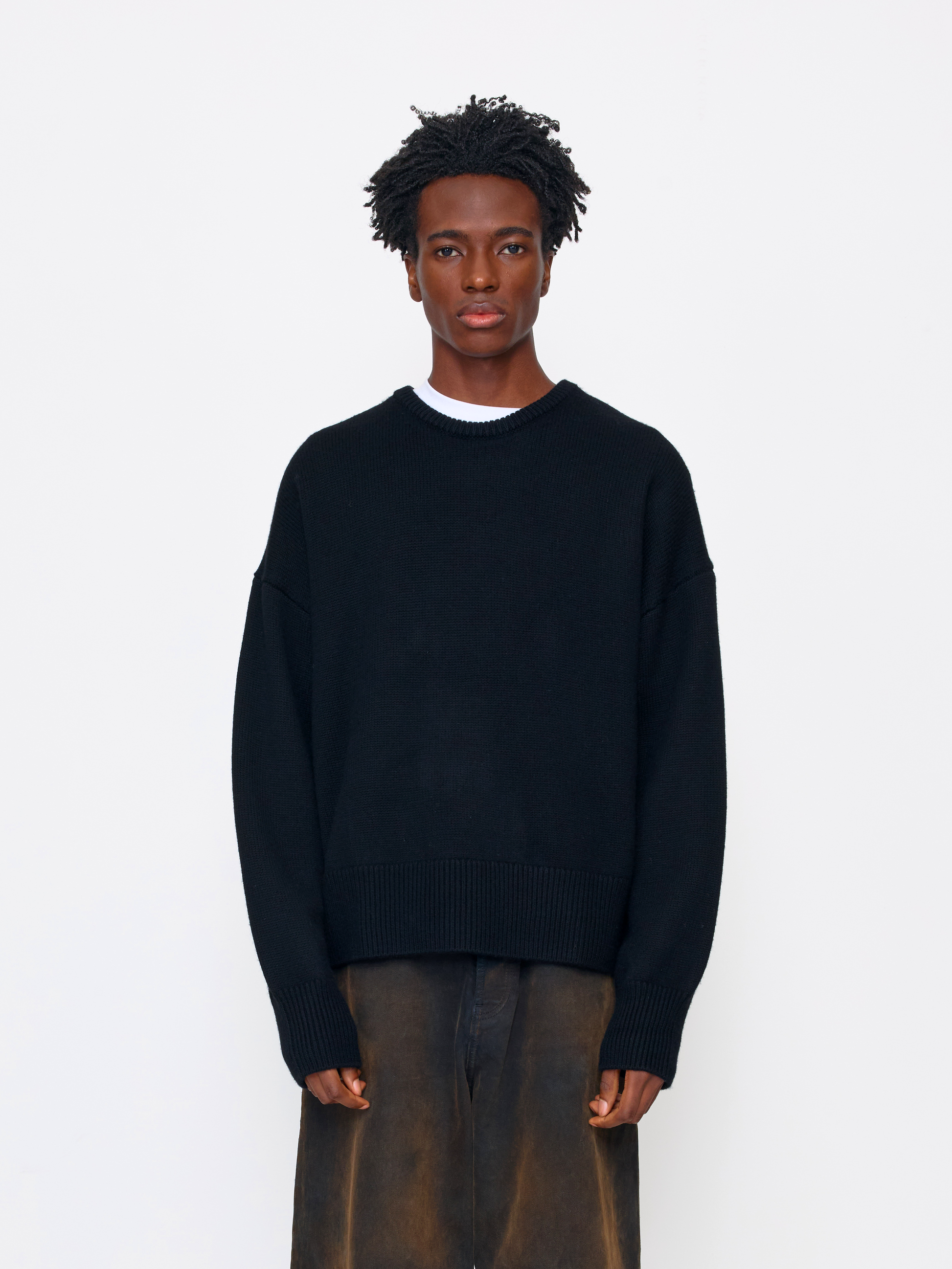 A model wearing a black knit crewneck sweater over a white undershirt and brown trousers, standing against a plain white studio background.